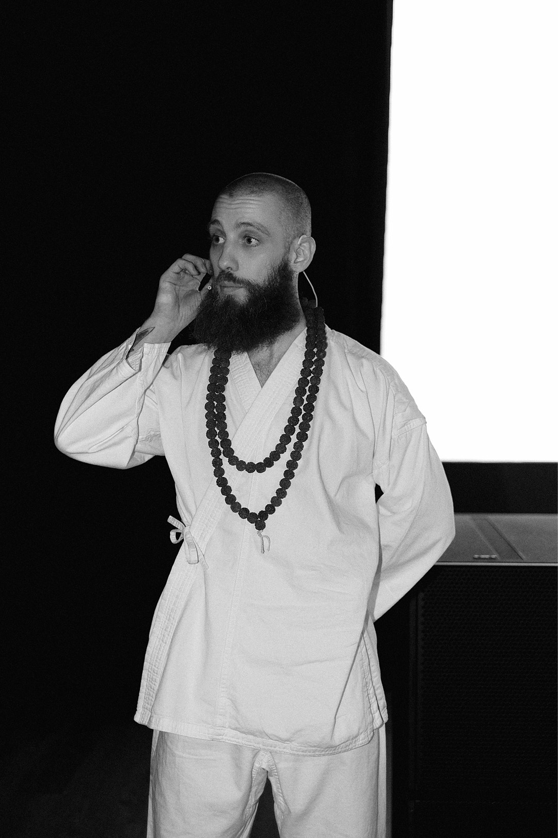 A man with a beard and shaved head wearing a white martial arts uniform and multiple beaded necklaces, standing indoors with a dark background and a large white screen or wall behind him, holding a headset or microphone near his ear.