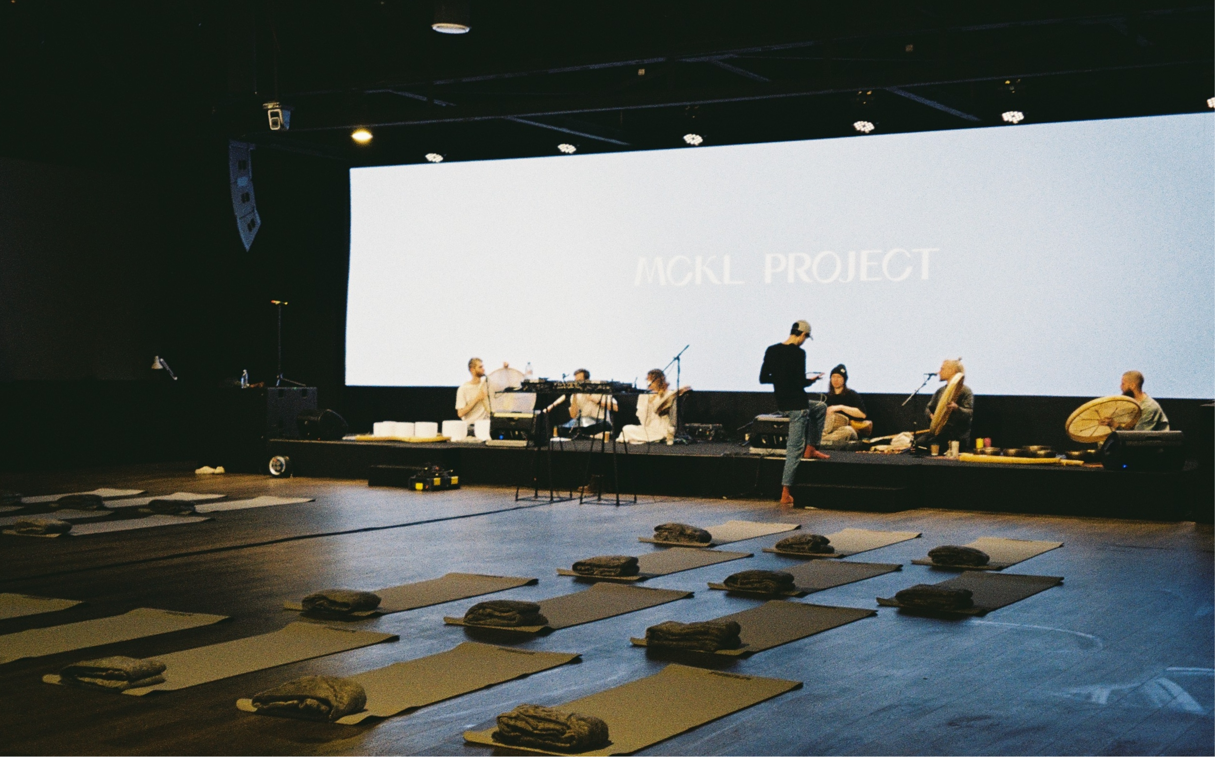 A music performance on stage with a large screen behind it displaying the words 'MCKL PROJECT'. There are several people on stage, some sitting and some standing, with musical instruments and equipment. In the foreground, there are mats and folded blankets arranged on the floor, suggesting a meditation or yoga session setup.
