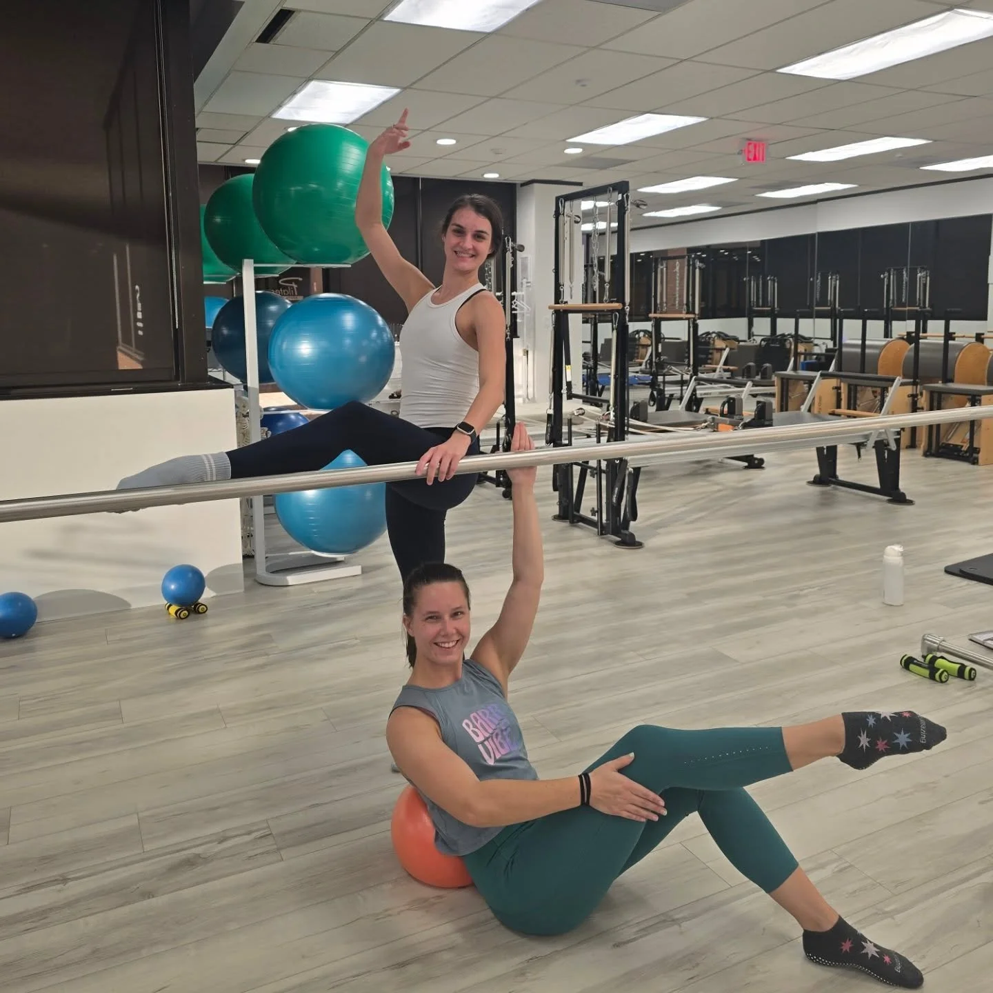 Two women in a gym performing an acro yoga pose. One is lying on the floor with a small exercise ball behind her back, supporting the other woman who is balancing on her hand and foot on a barre, smiling.