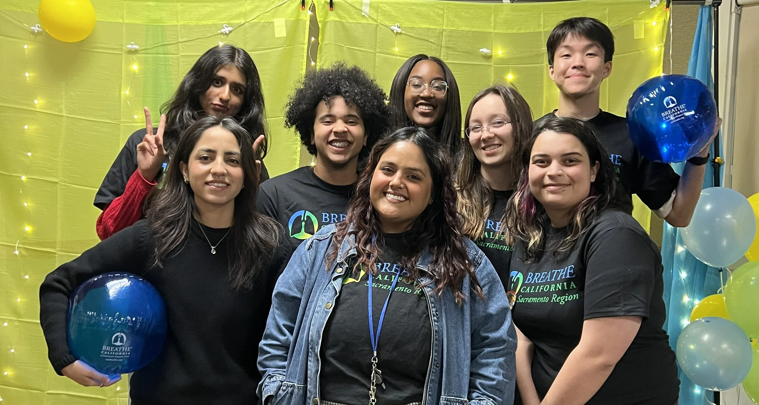 Six students standing together in front of a colorful backdrop, dressed casually, some wearing accessories like feather boas and a tiara, smiling for the camera.