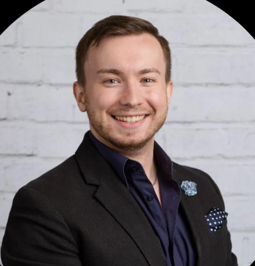 Portrait of a young man with short brown hair and a beard, wearing a dark suit jacket and navy shirt, smiling with a white brick wall background.