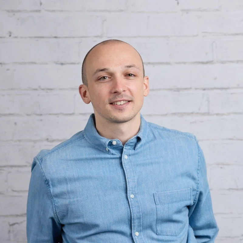 Portrait of a young man with a bald head, light skin, wearing a light blue button-up shirt, standing in front of a white brick wall.