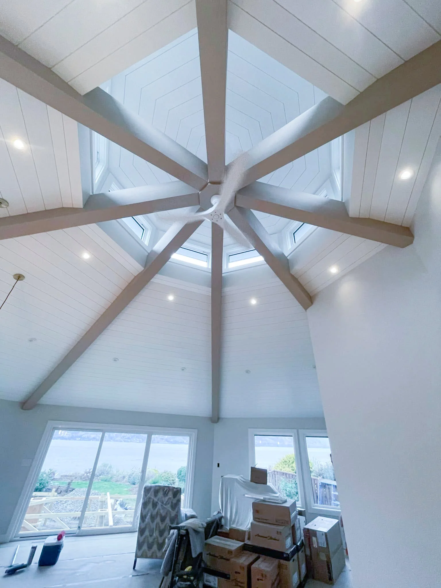 A room with a high vaulted ceiling and a central ceiling fan, featuring a large skylight at the top. The room has large windows with a view of the outdoors, where boxes and furniture are stored or being moved.