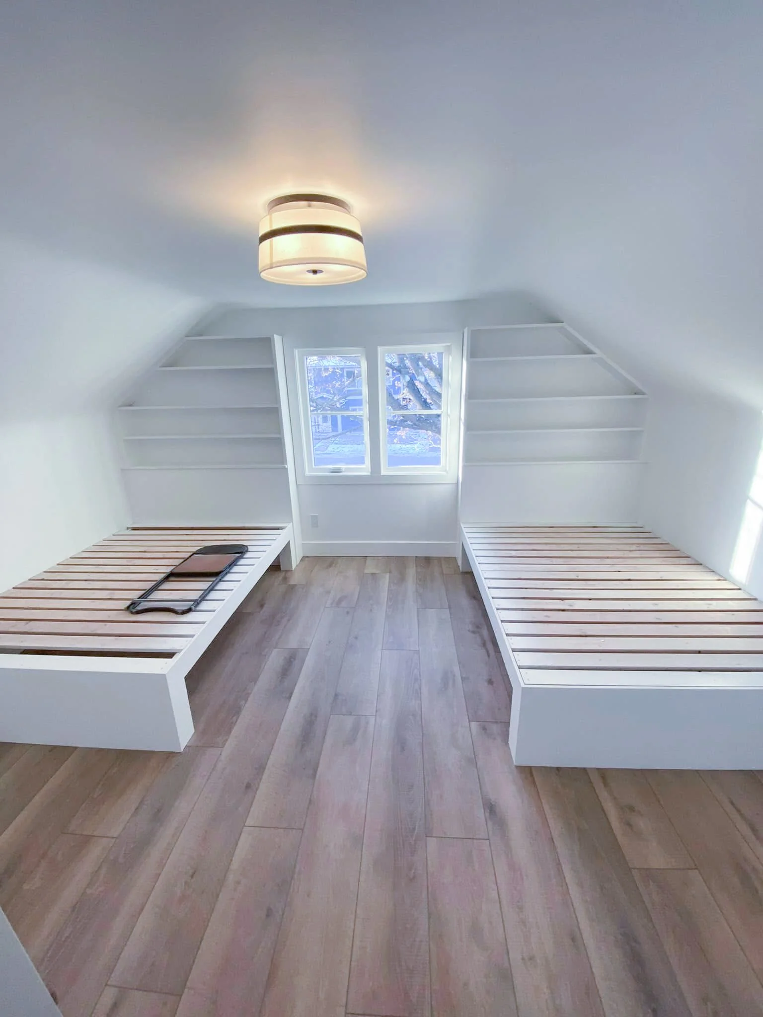 Room with white built-in beds and shelves, window, wood flooring, and ceiling light.