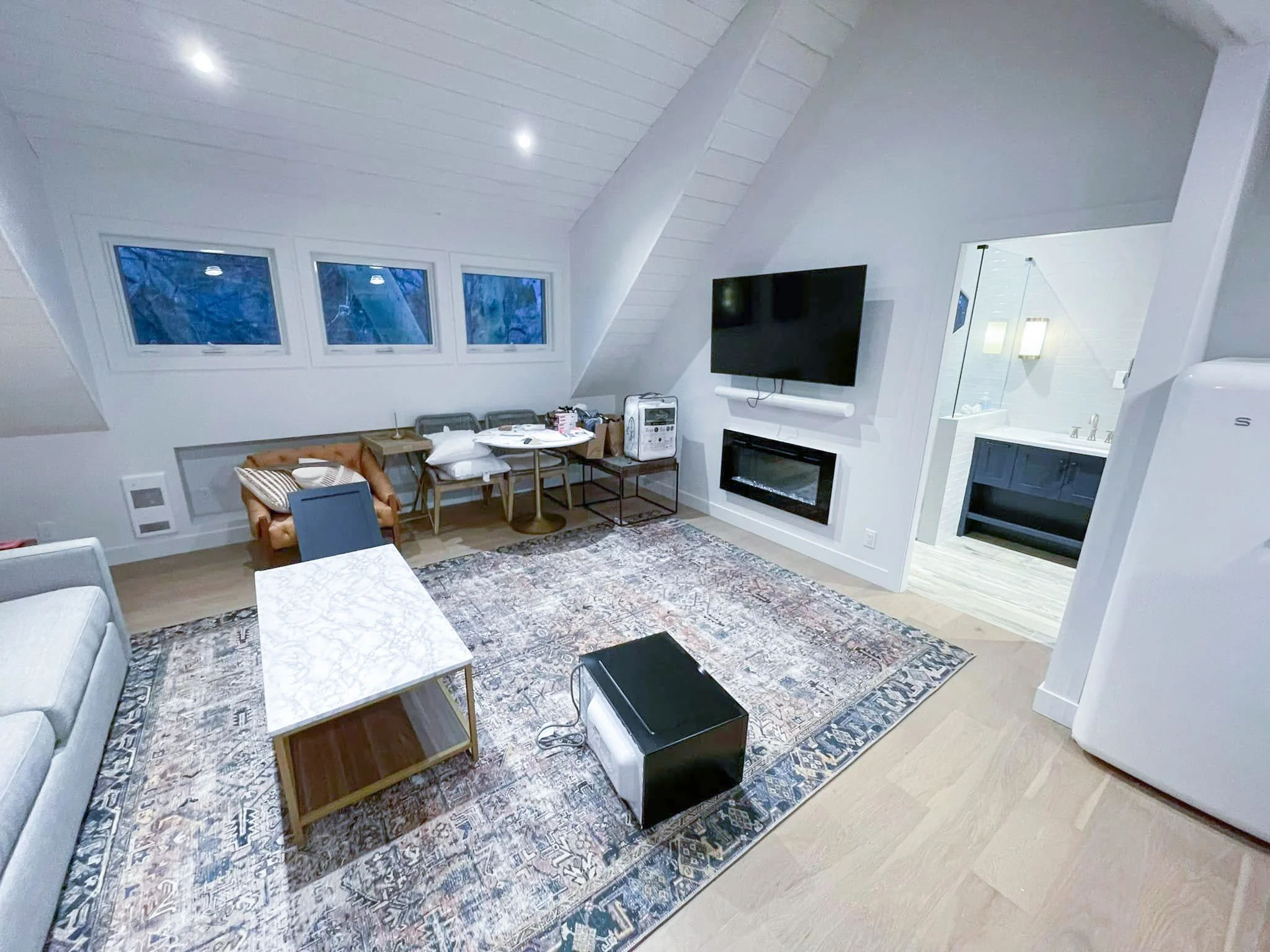 Living room with an area rug, a sofa, a coffee table, a large wall-mounted TV, a fireplace, and windows showing a blue sky outside.