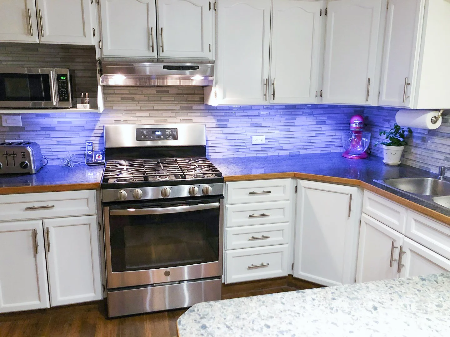 Kitchen with white cabinets, dark countertops, stainless steel stove, microwave, toaster, water kettle, potted plant, pink stand mixer, paper towel roll, and blue LED lighting under the cabinets.