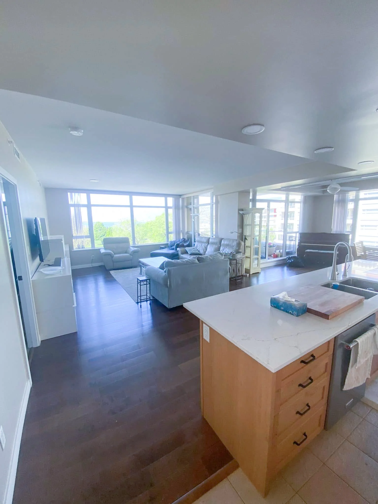 Open-concept living room with large windows, light-colored furniture, and hardwood flooring, viewed from the kitchen.
