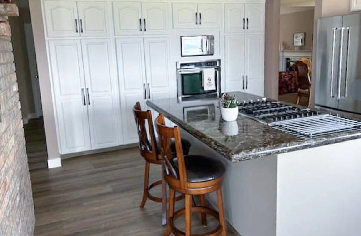 Modern kitchen with white cabinets, a granite island with two wooden barstools, stainless steel microwave, oven, and refrigerator, and hardwood floors.