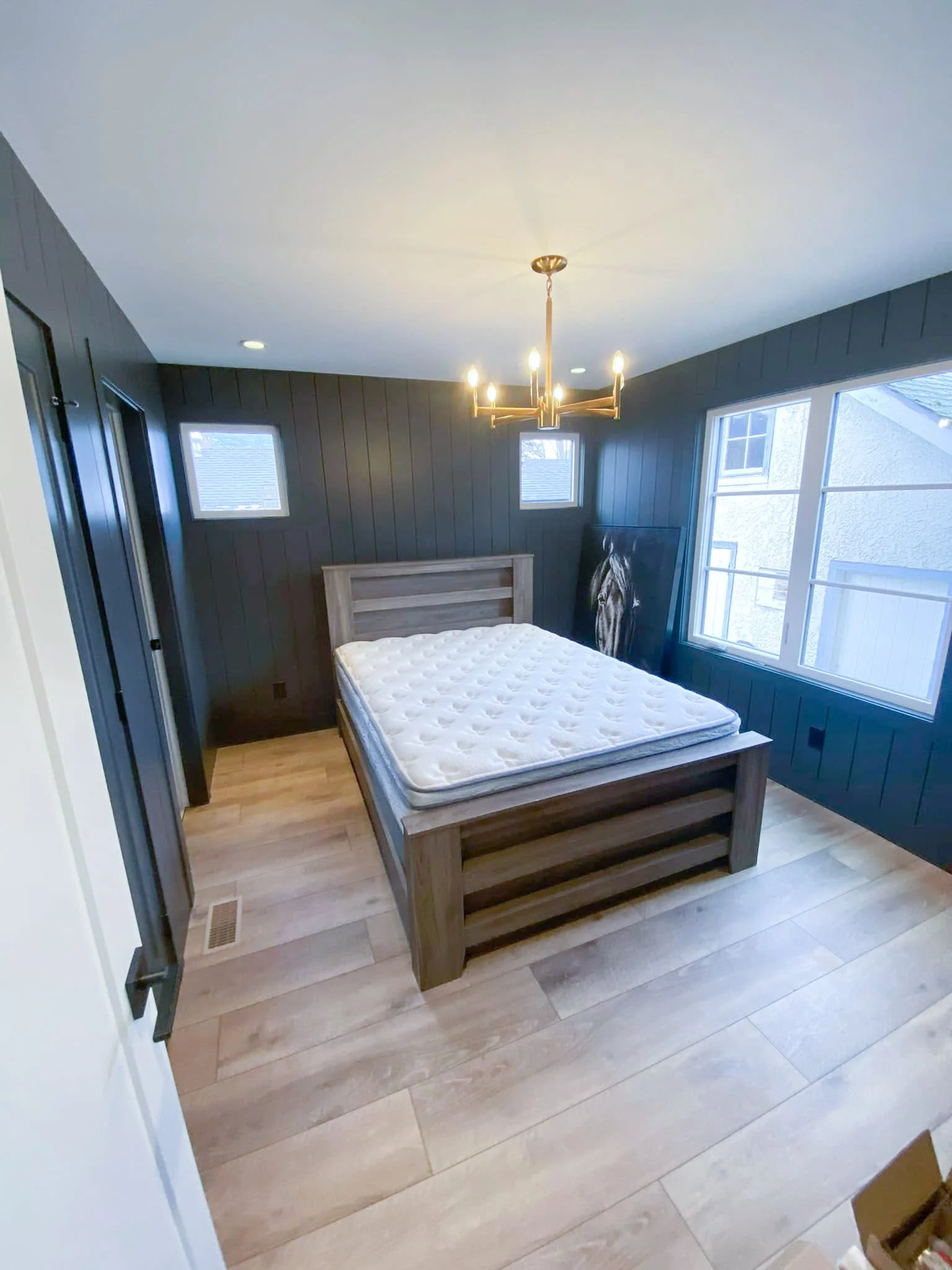 Empty bedroom with dark paneled walls, a large window, a chandelier, and a bed with a mattress but no bedding, featuring wooden flooring and a painting leaning against the wall.
