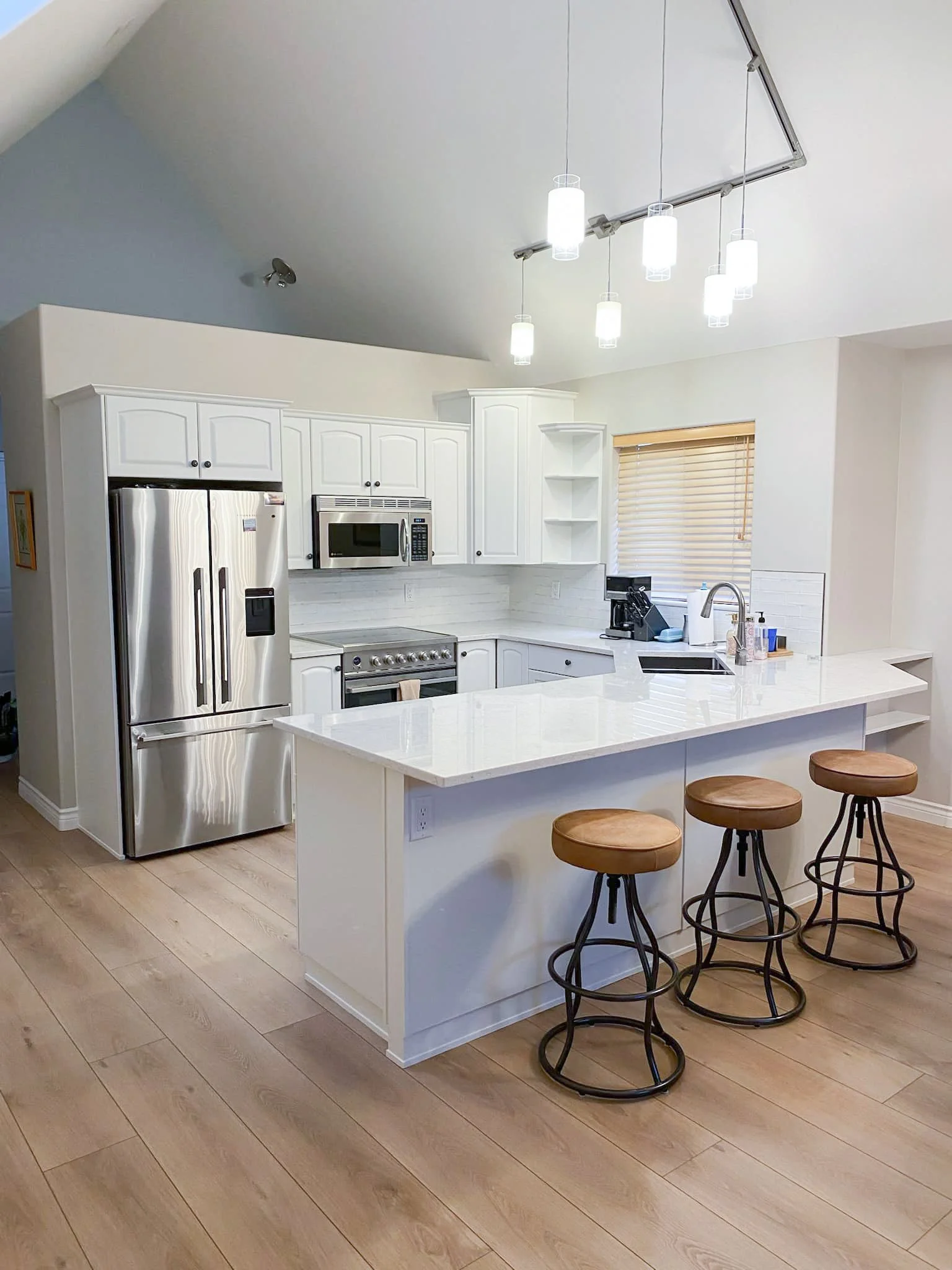 Modern white kitchen with island, three bar stools, stainless steel refrigerator, microwave, stove, and coffee maker, under a sloped ceiling with hanging lights.