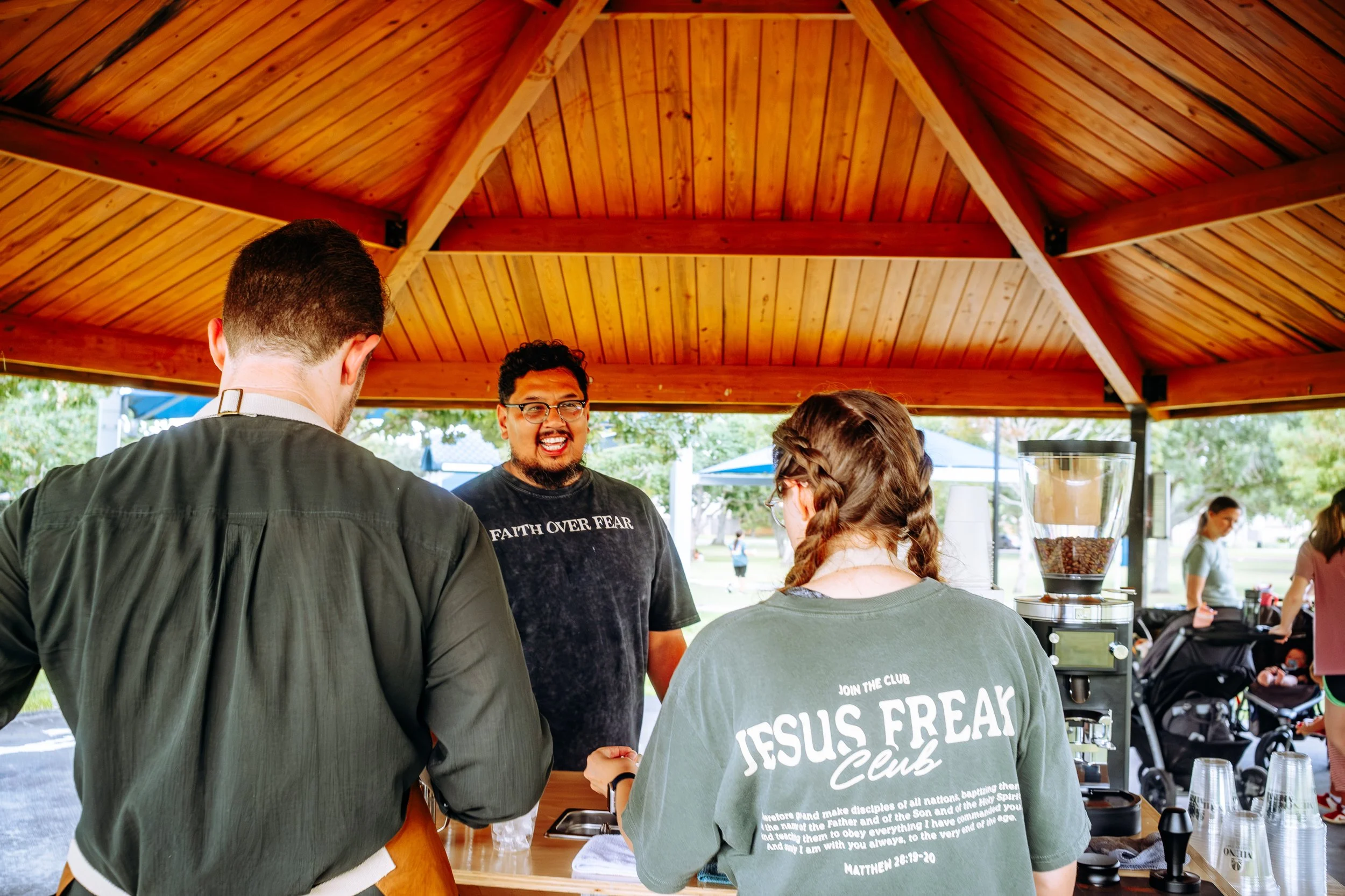 People at an outdoor coffee stand at a park, with a man smiling and wearing a black t-shirt that says 'Faith Over Fear' and a woman with braided hair Coffee Catering in Houston, Texas Coffee Cart and Mobile Espresso Bar Event Baristas. Coffee Vendor