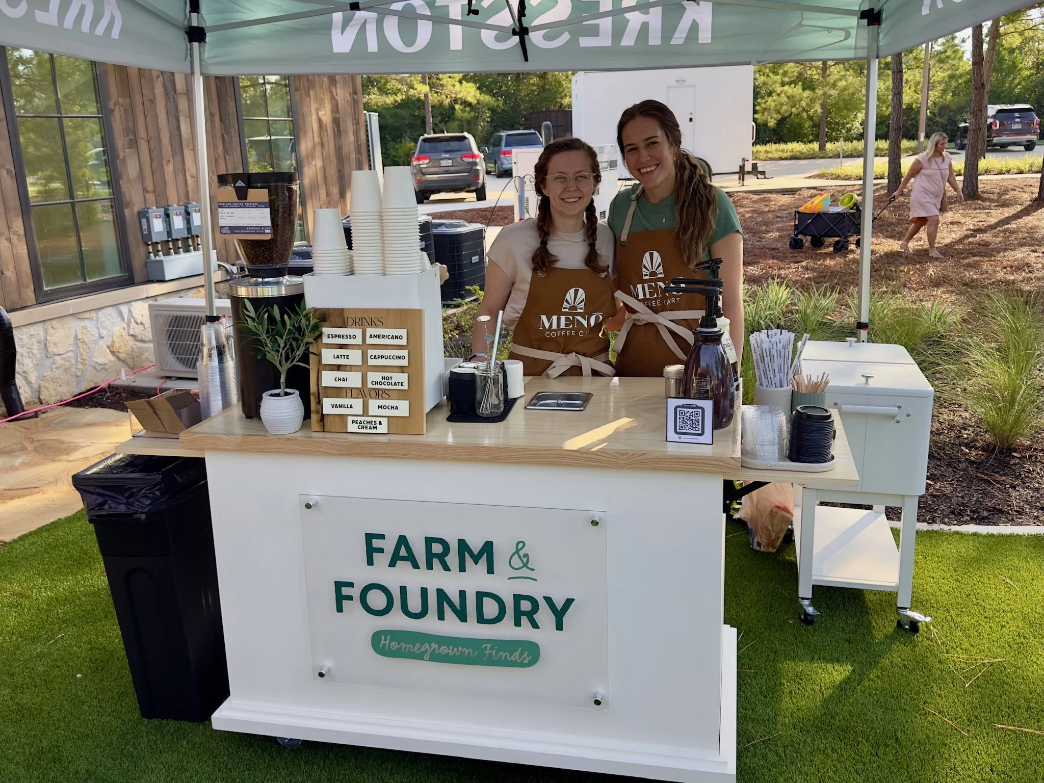 Two women smiling at Meno coffee cart labeled 'Farm & Foundry Homegrown Finds' under a canopy, with coffee cups, a menu board, and a QR code on the counter.