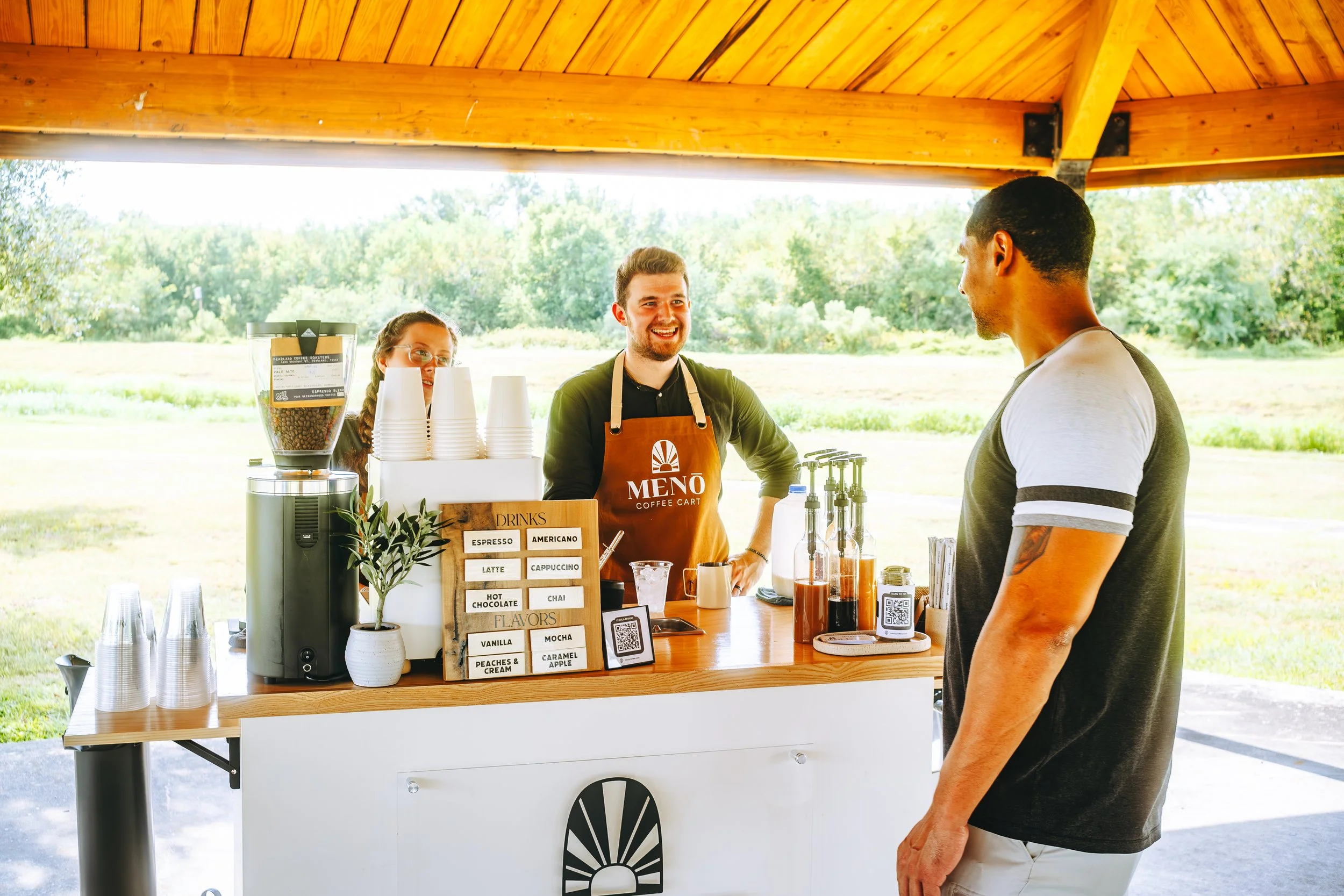 A coffee stand with a barista serving a customer outdoors on a sunny day, with trees and grass in the background. Coffee Catering in Houston, Texas Coffee Cart and Mobile Espresso Bar Event Baristas. Coffee Vendor services