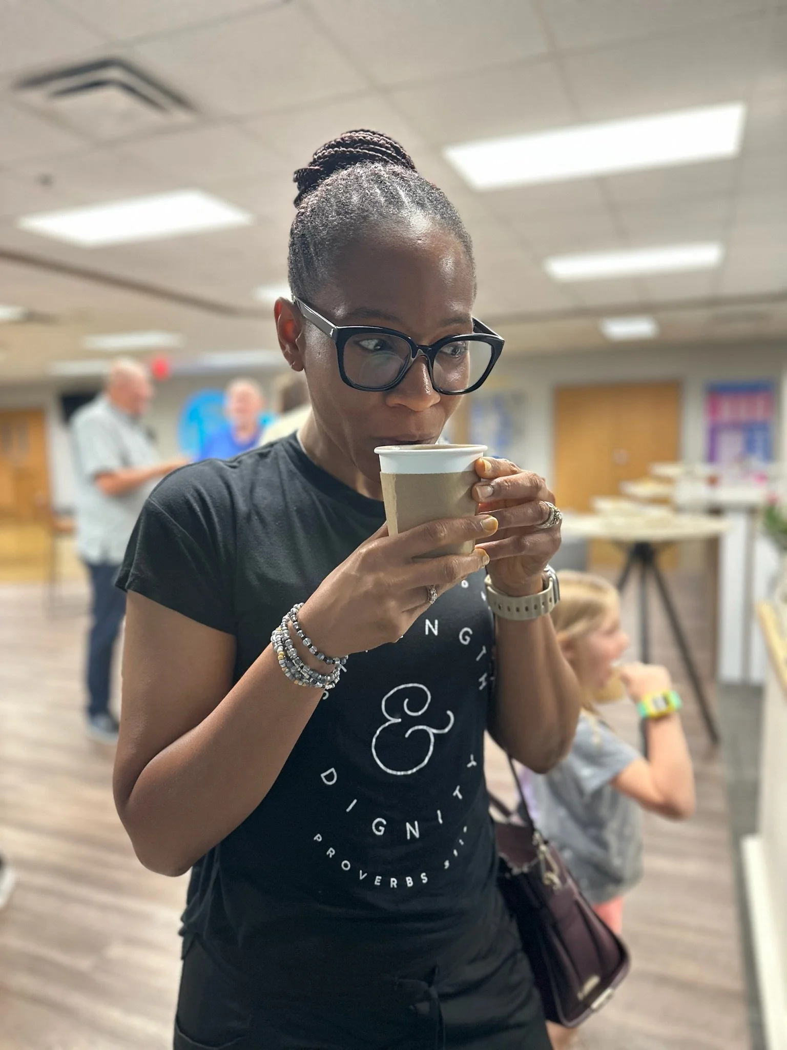 A woman wearing glasses and a black t-shirt sipping from a paper coffee cup in an indoor setting.