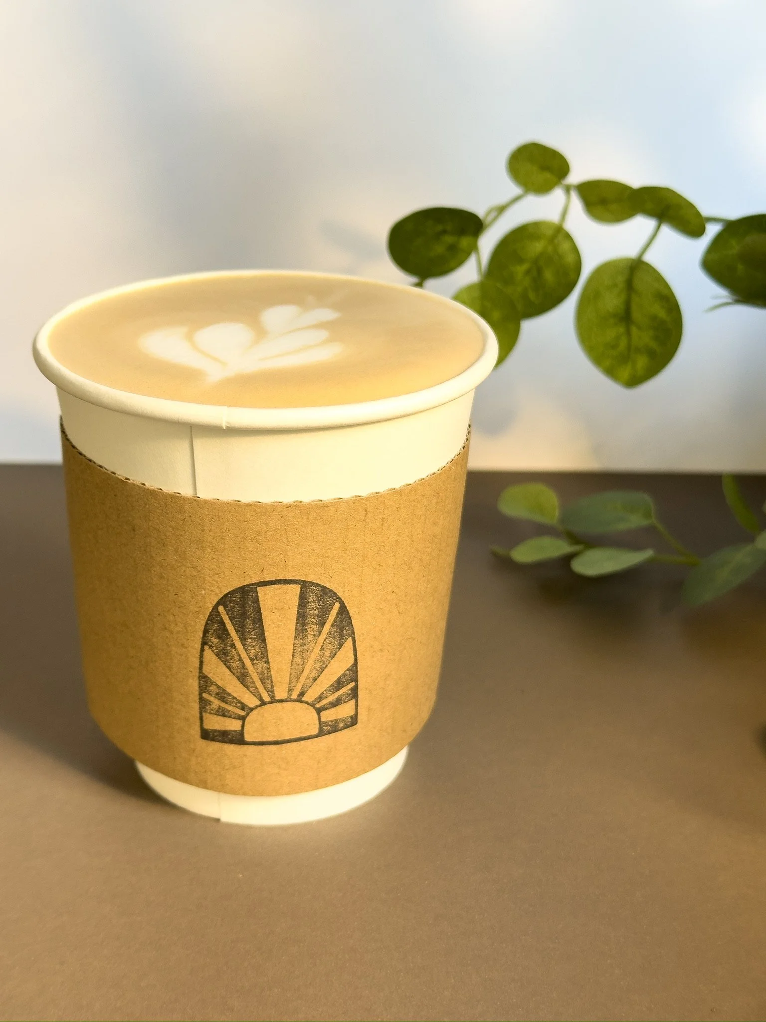 Custom Coffee Cup with a cardboard sleeve, placed on a gray surface with a green leafy plant in the background. Coffee Catering in Houston, Texas Coffee Cart and Mobile Espresso Bar Event Baristas. Coffee Vendor Services