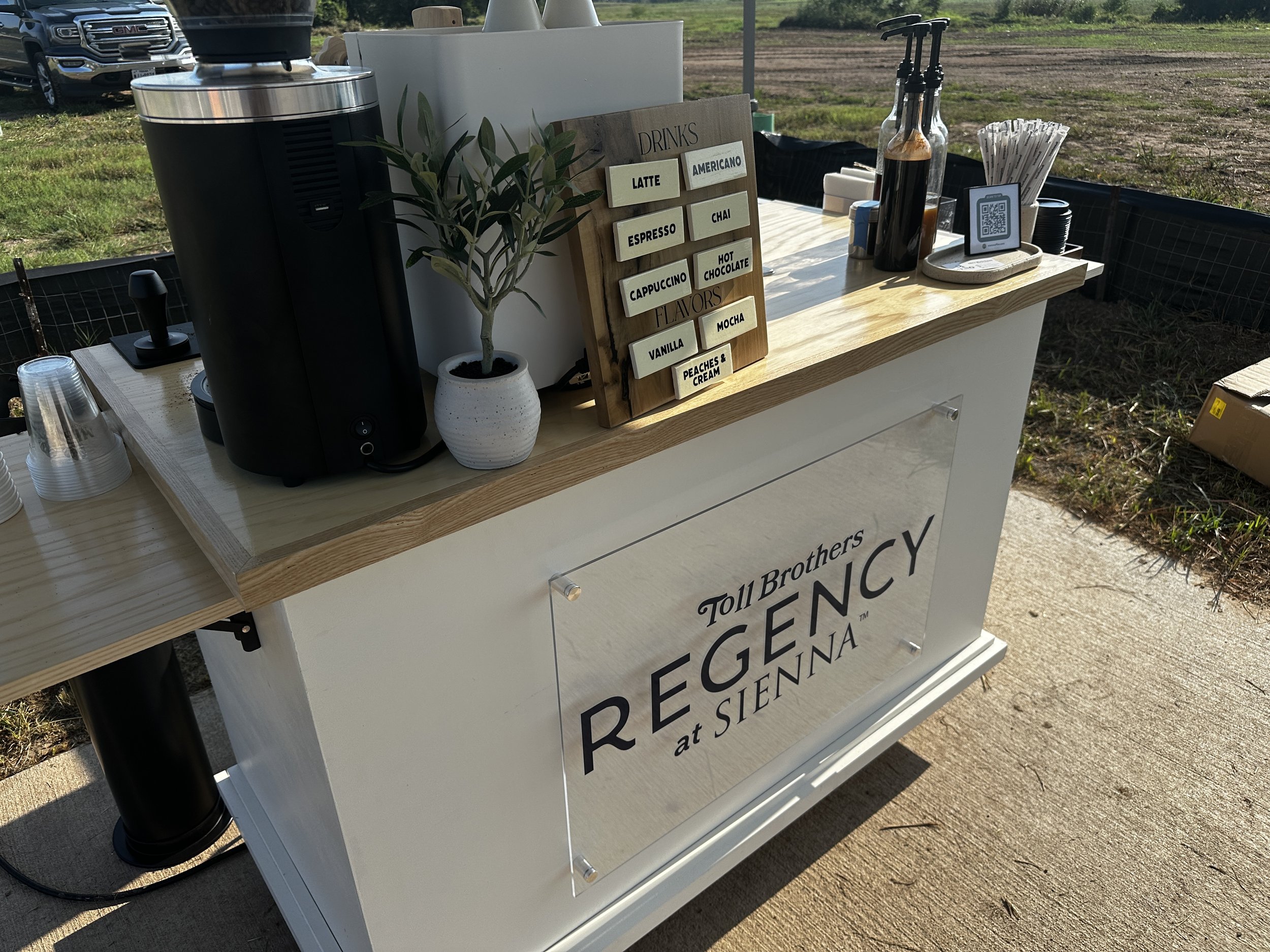 Coffee and beverage station at an outdoor event with a sign for Toll Brothers Regency at Sienna. Coffee Catering in Houston, Texas Coffee Cart and Mobile Espresso Bar Event Baristas. Coffee Vendor Services