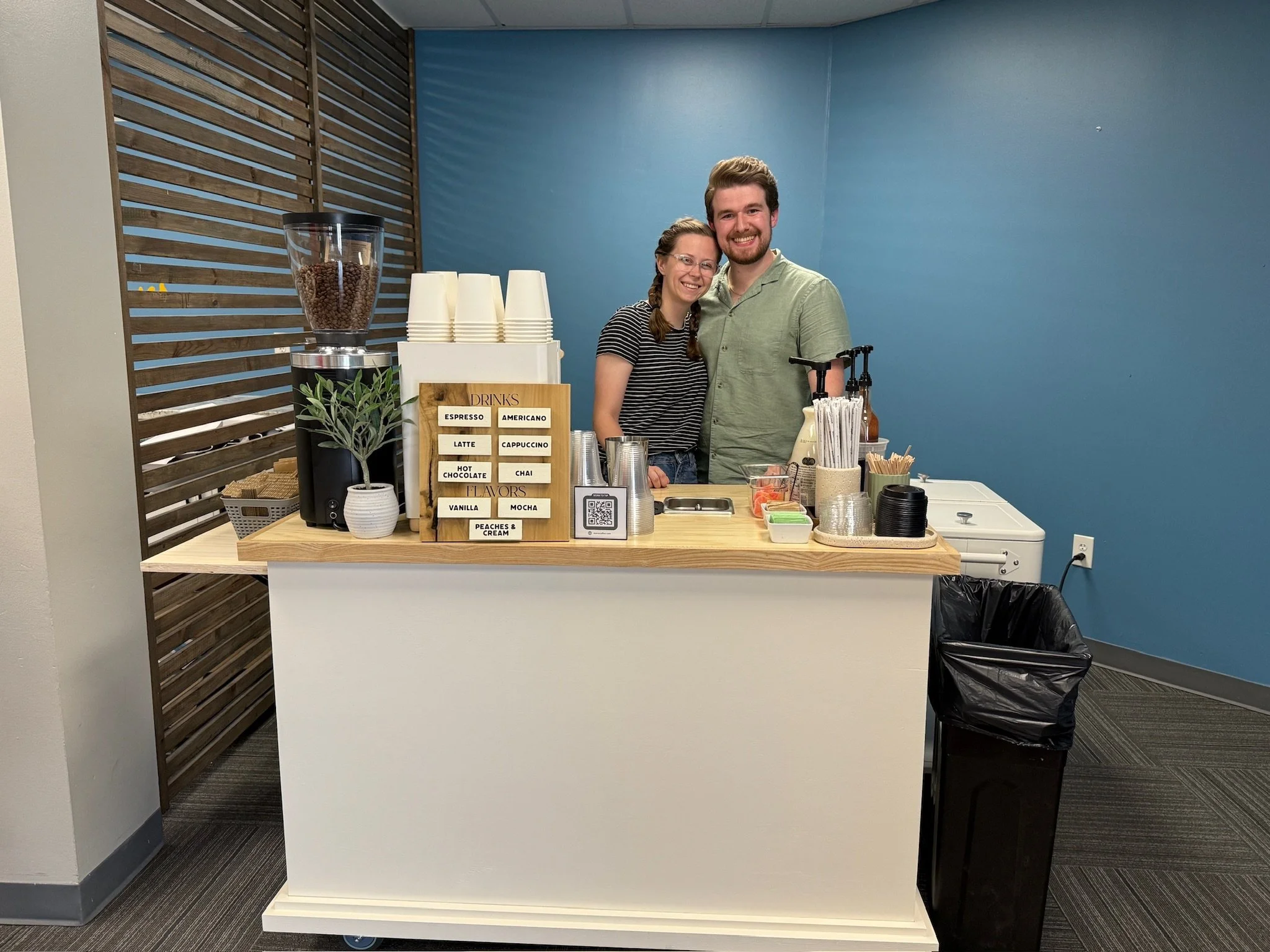 A smiling man and woman standing behind Meno coffee cart in a church. The stand has a coffee grinder, cups, and drink flavor menu, with a trash bin beside it.