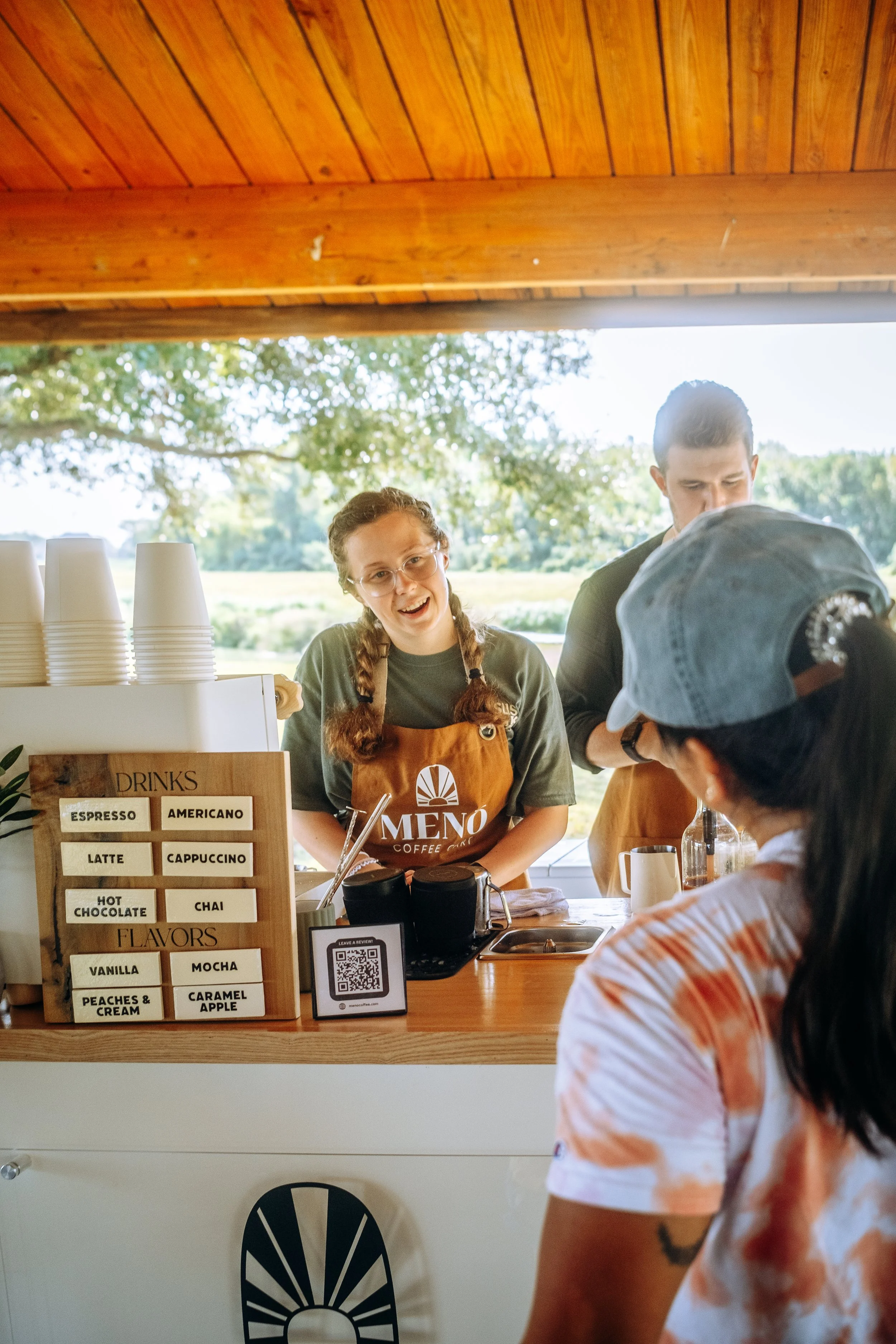 A woman at a coffee stand smiling and talking to a customer, with two other customers in front of her, and a man standing nearby. Coffee Catering in Houston, Texas Coffee Cart and Mobile Espresso Bar Event Baristas. Coffee Vendor services
