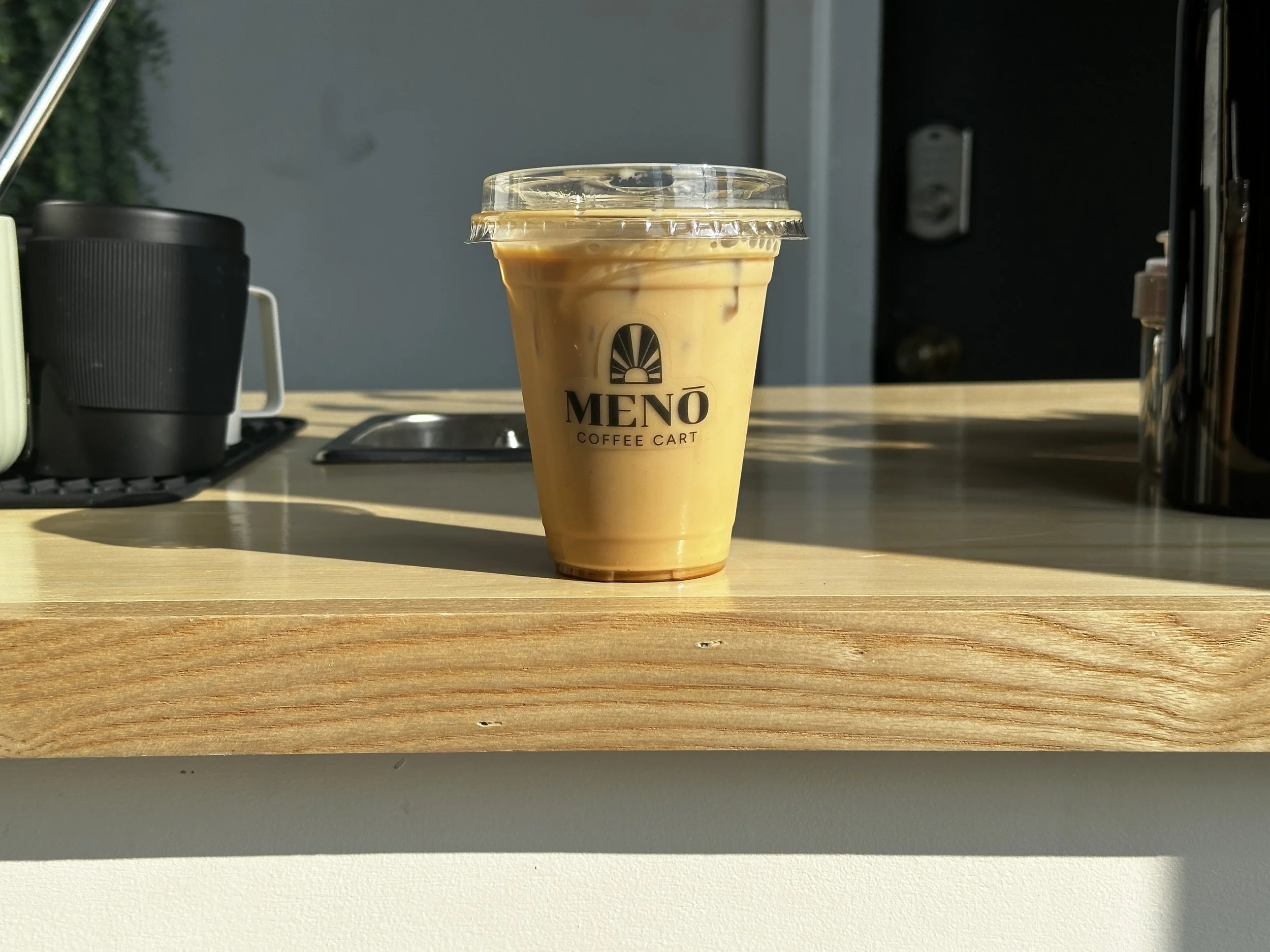 Iced coffee in a clear plastic cup with a black and gold logo reading "MENO Coffee Cart" Coffee Catering in Houston, Texas Coffee Cart and Mobile Espresso Bar Event Baristas. Coffee Vendor Services