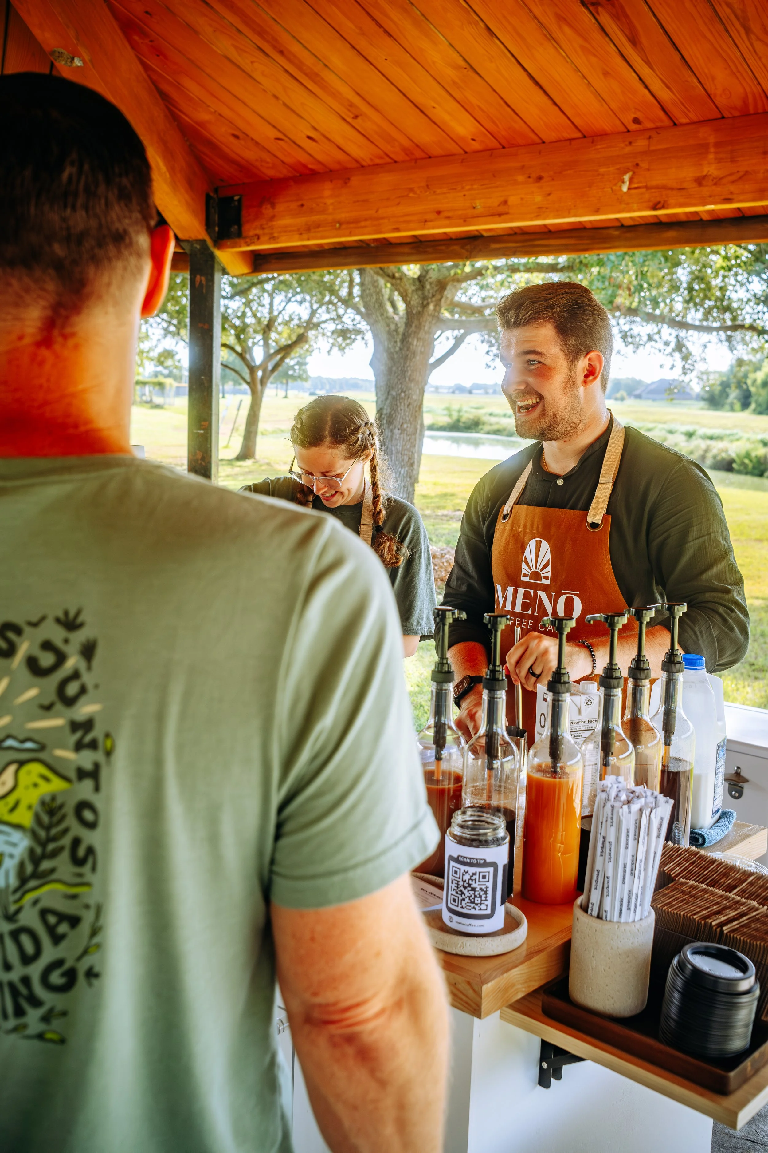 A cheerful barista serving drinks at an outdoor coffee stand with a scenic park and trees in the background. Coffee Catering in Houston, Texas Coffee Cart and Mobile Espresso Bar Event Baristas. Coffee Vendor services