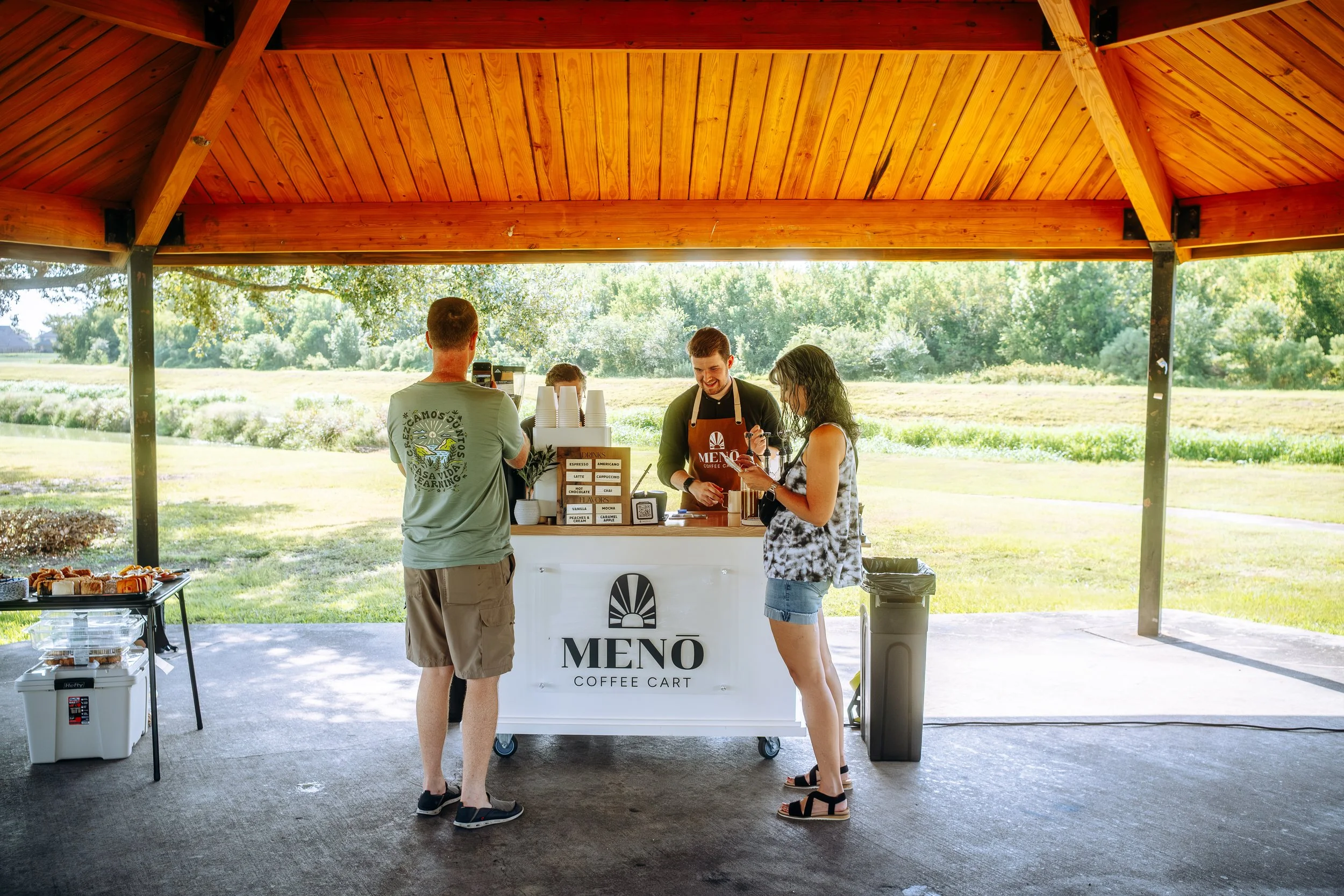 A small outdoor coffee stand called 'MENÖ Coffee Cart' with a man and woman ordering drinks from a barista under a wooden pavilion, Coffee Catering in Houston, Texas Coffee Cart and Mobile Espresso Bar Event Baristas. Coffee Vendor services