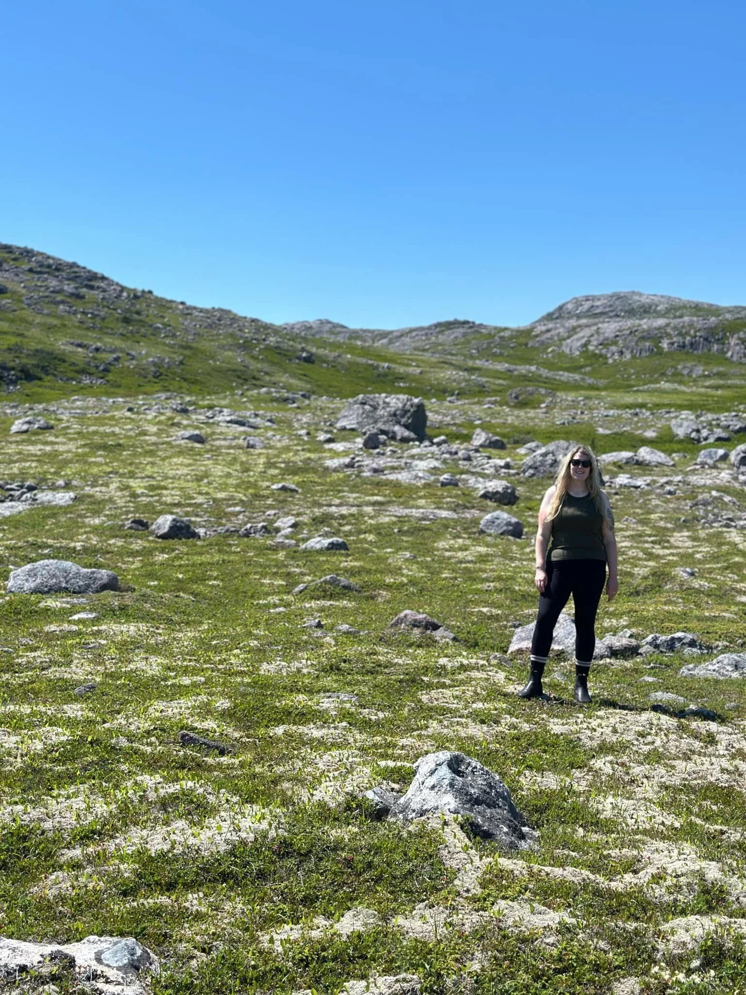 Woman standing in a rocky green landscape in Saint Pierre and Miquelon under a clear blue sky.