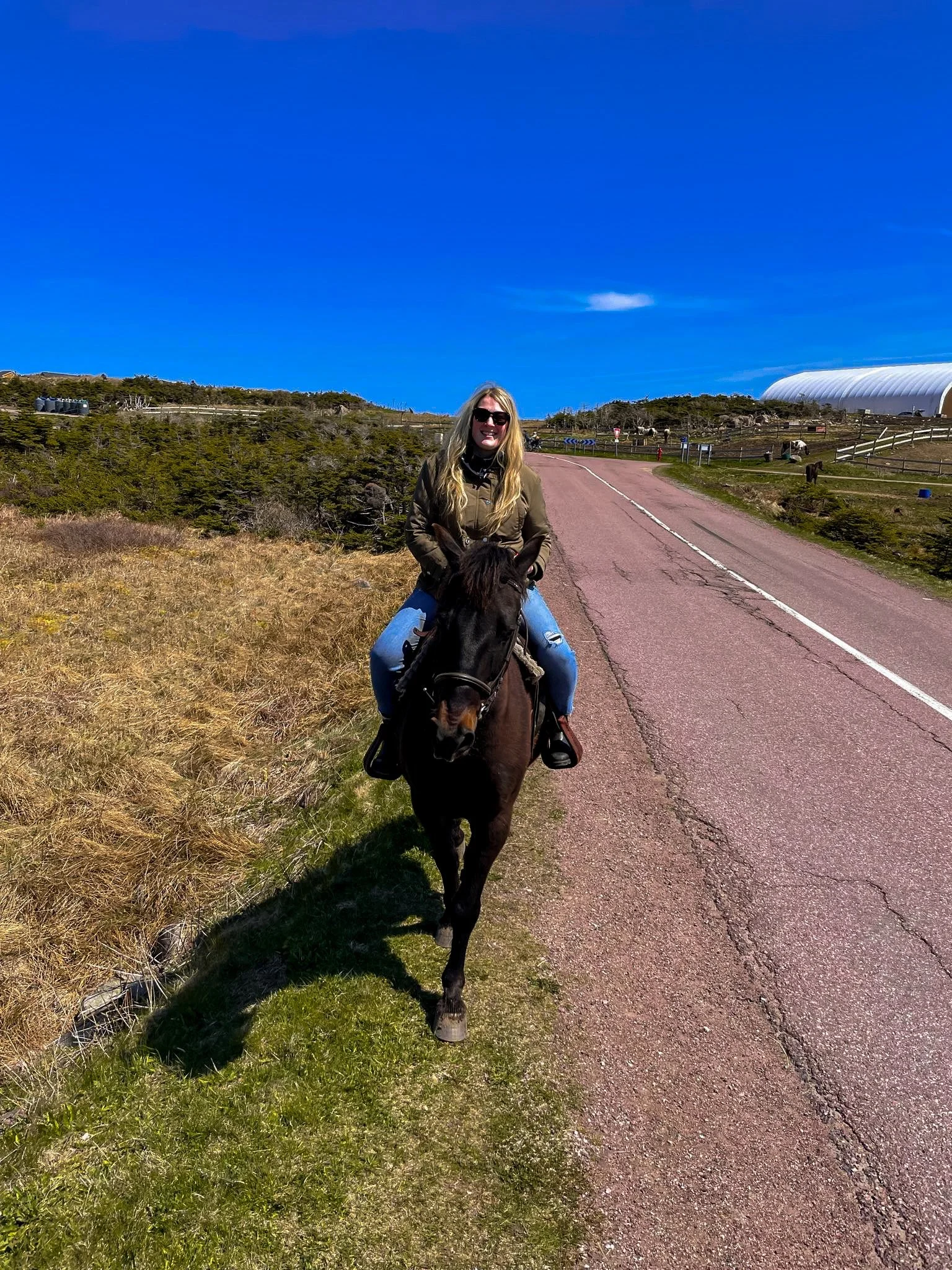 Woman riding a horse along a rural road in Saint Pierre and Miquelon under a bright blue sky.