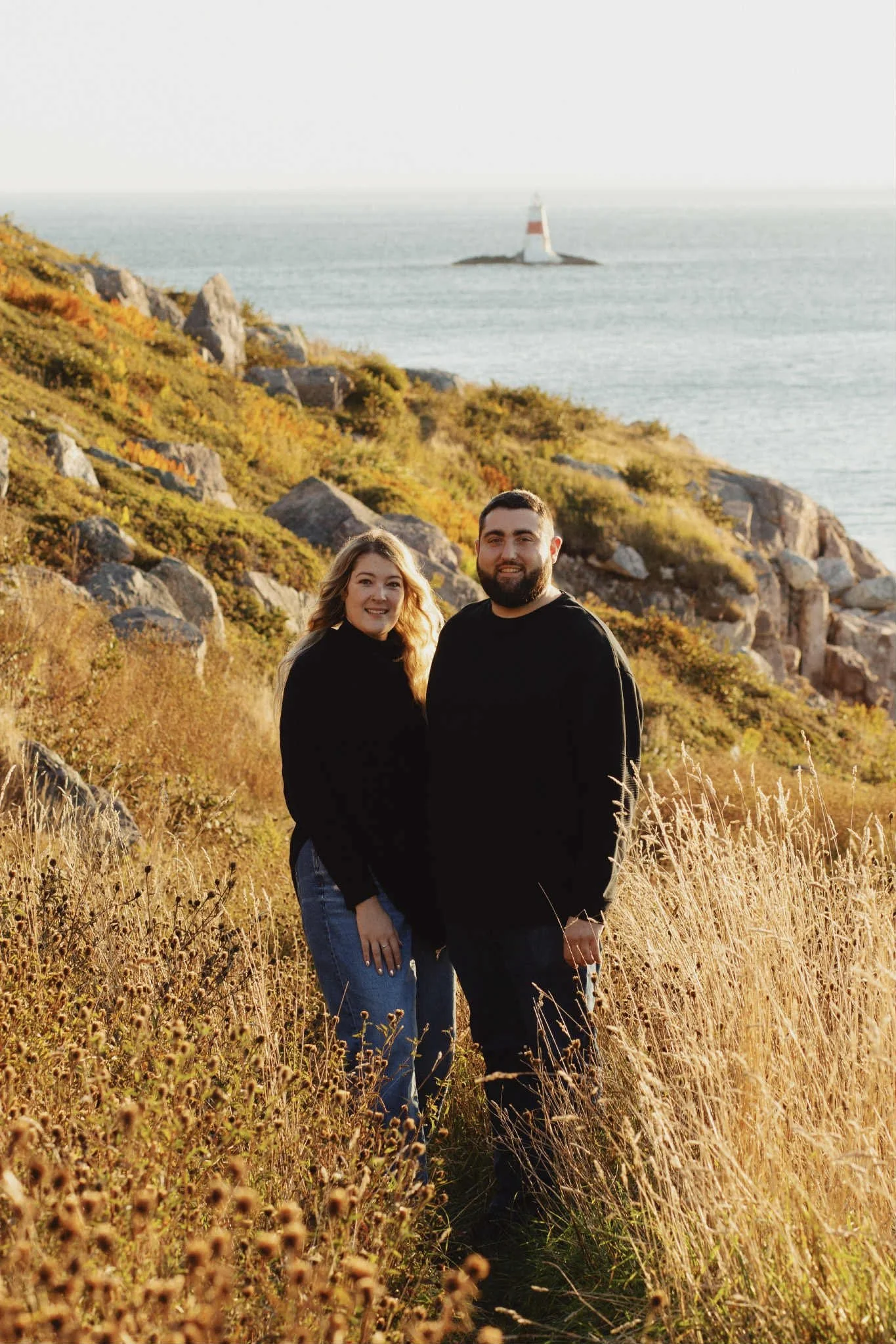 Couple standing on a coastal trail in Saint Pierre and Miquelon with rocky cliffs and a lighthouse in the background.