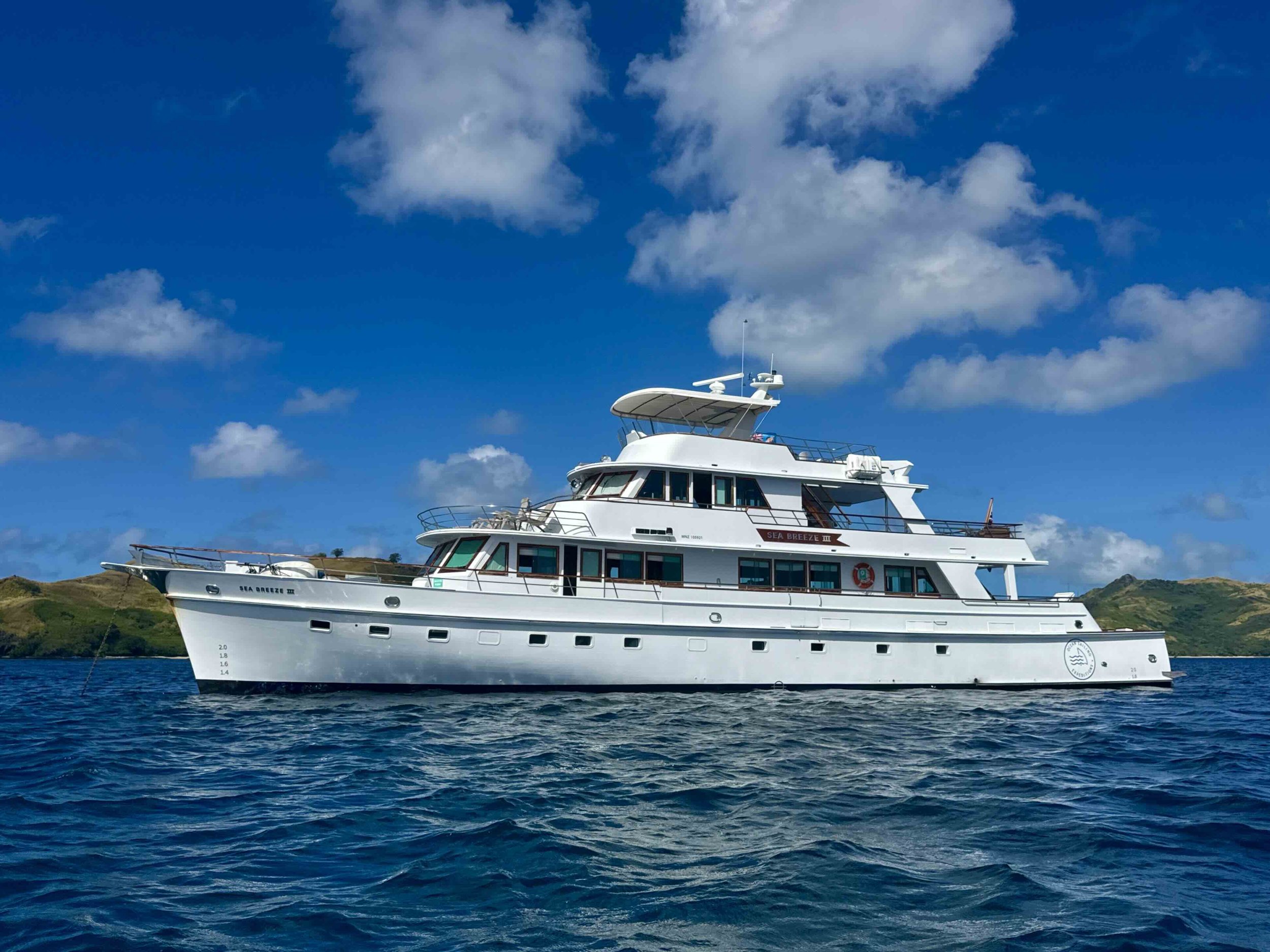 A white yacht named Sea Breeze III floating on the water with a green island in the background under a partly cloudy sky.