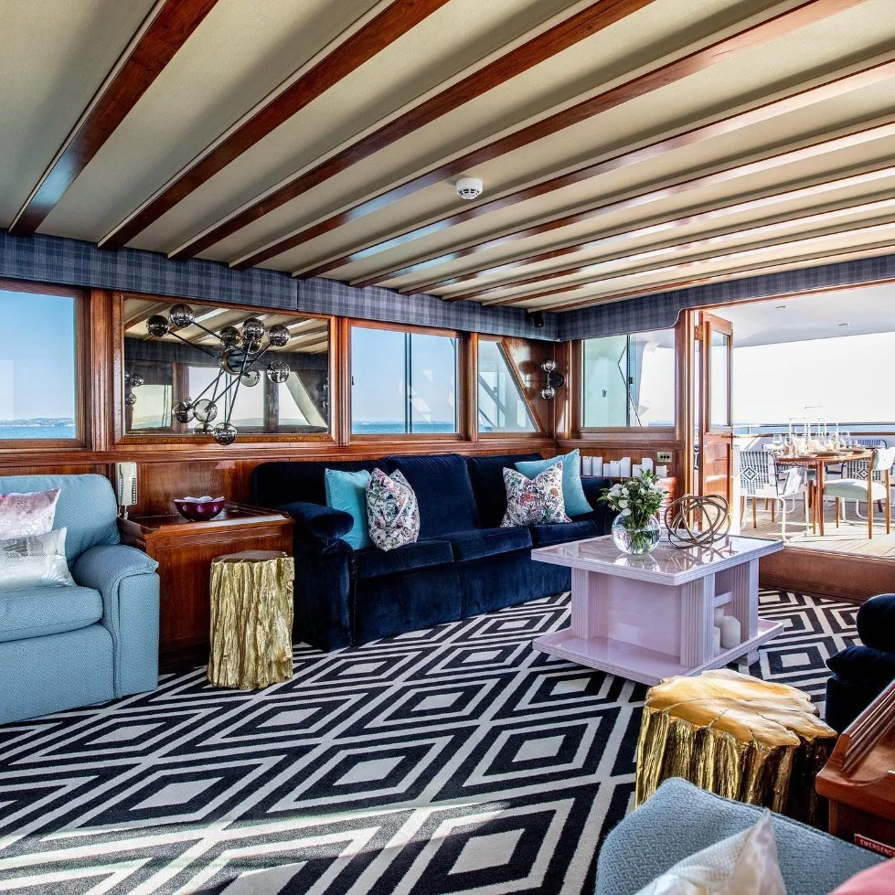 Interior view of a stylish living room on a boat or yacht with large windows showing an ocean view. The room features a black velvet sofa, a light blue armchair, and a pink coffee table with decorative items on it. The floor has a black and white geo