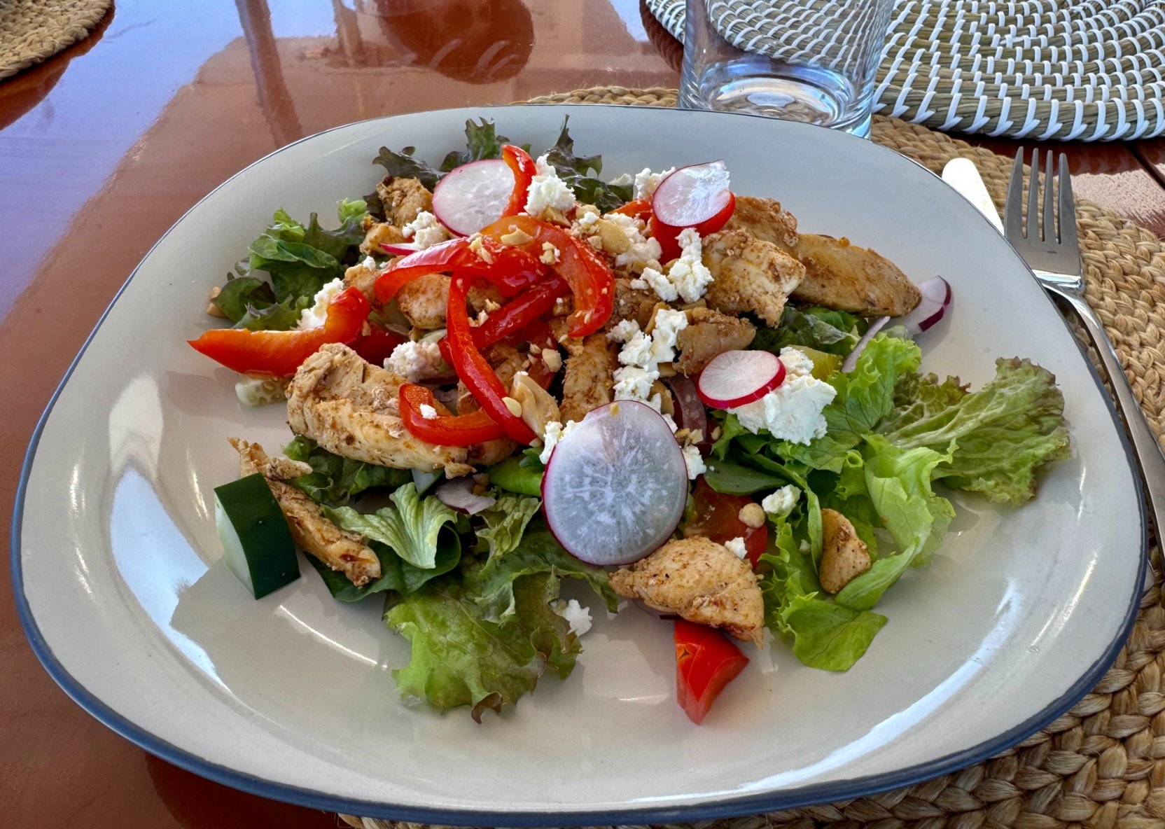 A plate of fresh salad with grilled chicken, mixed greens, sliced radishes, cherry tomatoes, red bell peppers, and crumbled cheese, on a woven placemat with a glass of water and cutlery nearby.