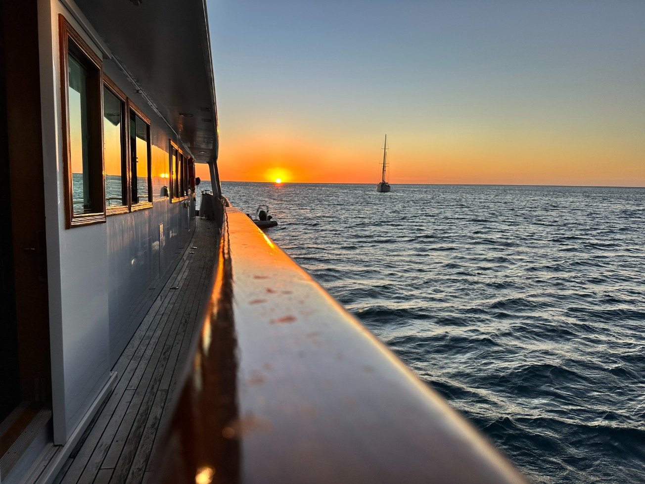 Sunset view from the deck of a boat, with a sailboat in the distance and the sky transitioning from orange to blue.