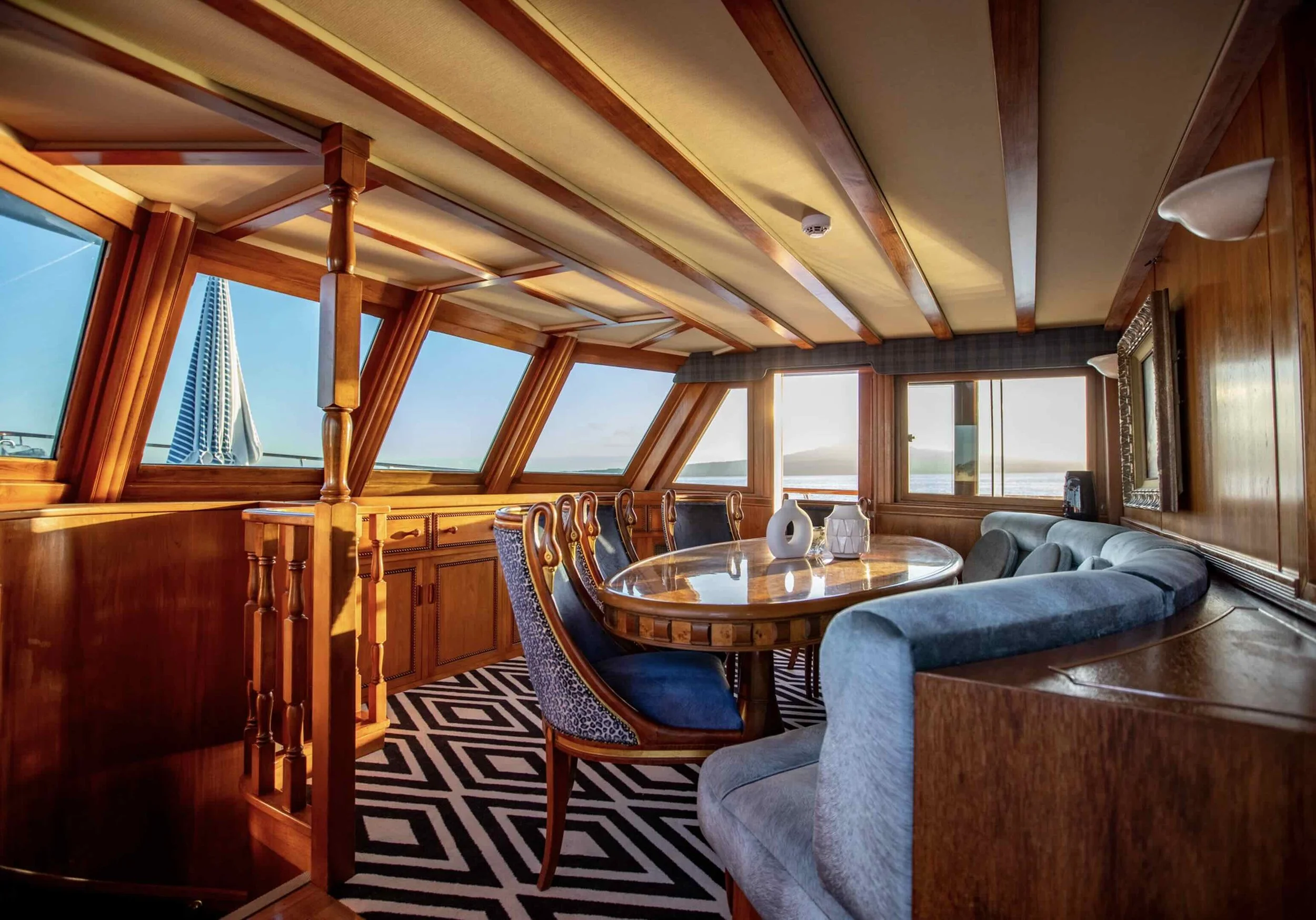 Interior of a yacht's dining area with wooden paneling, a round table, upholstered chairs, large windows showing the ocean and clear sky, and decorative vases on the table.