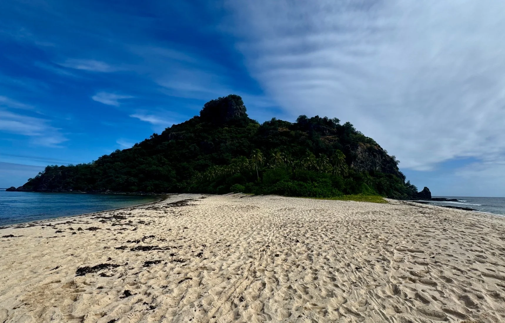 A tropical island with a sandy beach, green vegetation, and a hill or mountain, under a partly cloudy sky, with the ocean on both sides.