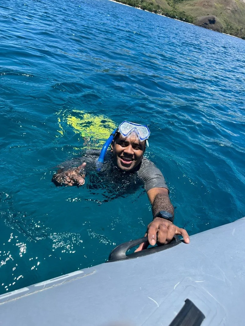 A man with snorkeling gear, smiling and reaching for a boat's side while in clear blue water, with green hills in the background.