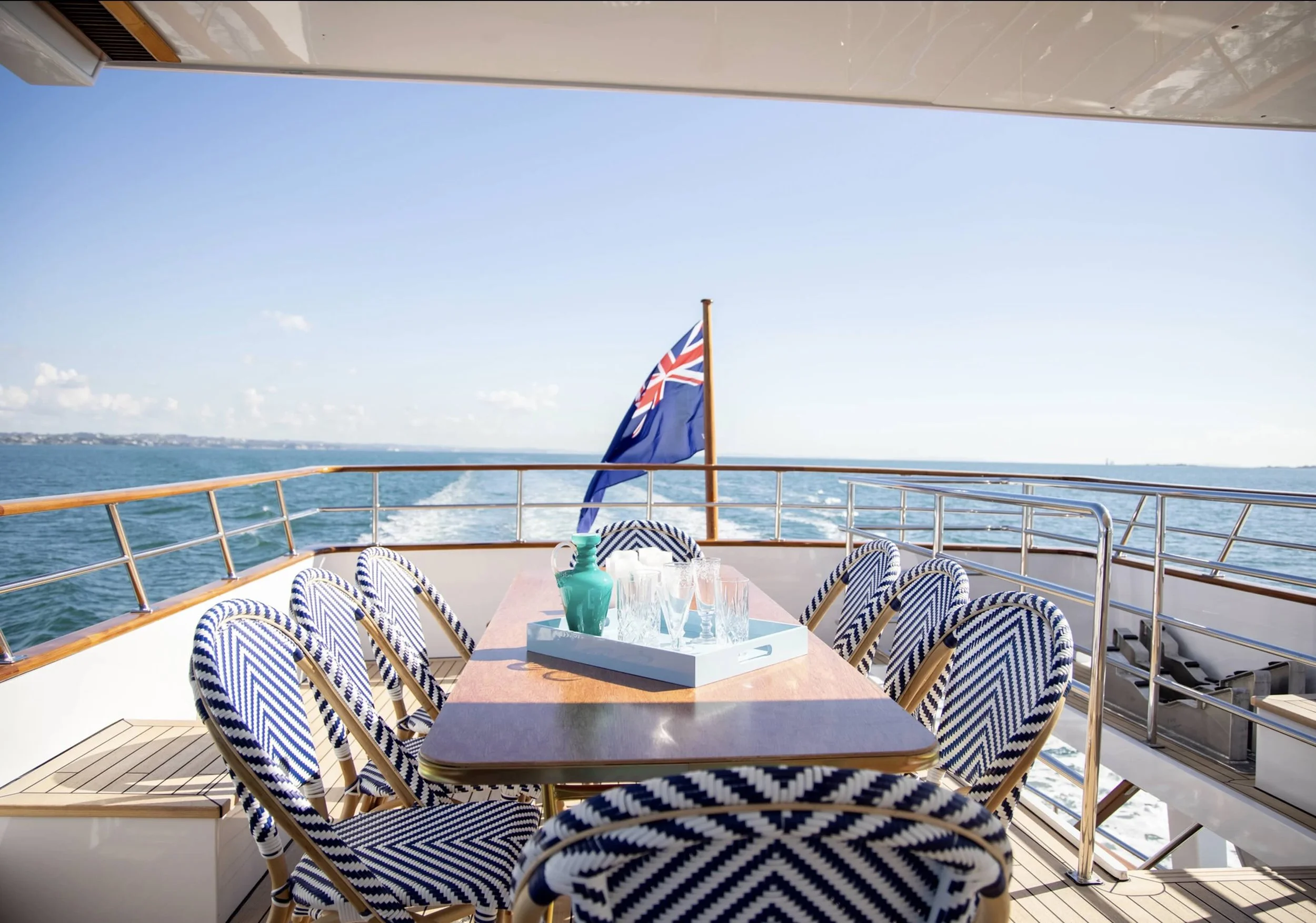 Outdoor dining area on a boat deck with a wooden table, blue and white patterned chairs, a tray with glasses and a green pitcher, railing, and an Australian flag in the background over the ocean with a distant shoreline and a clear sky.