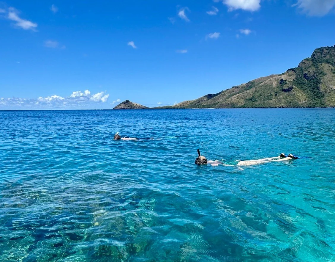 Three snorkelers swimming in clear blue ocean water near green islands and rocky hills under a partly cloudy sky.