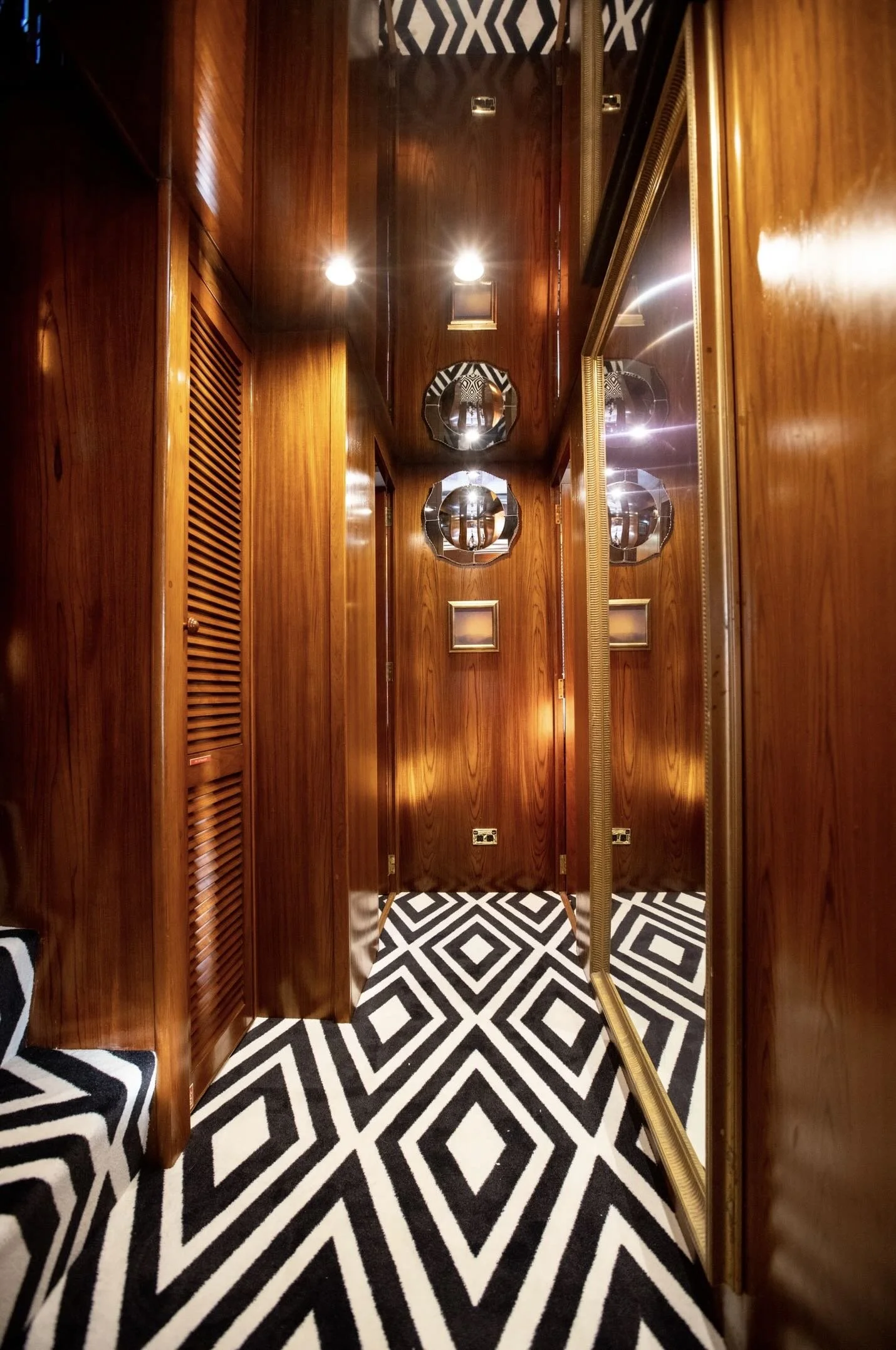 Interior of a wood-paneled room with a black and white geometric patterned carpet, mirror, and decorative mirrors on the walls, illuminated by ceiling lights.
