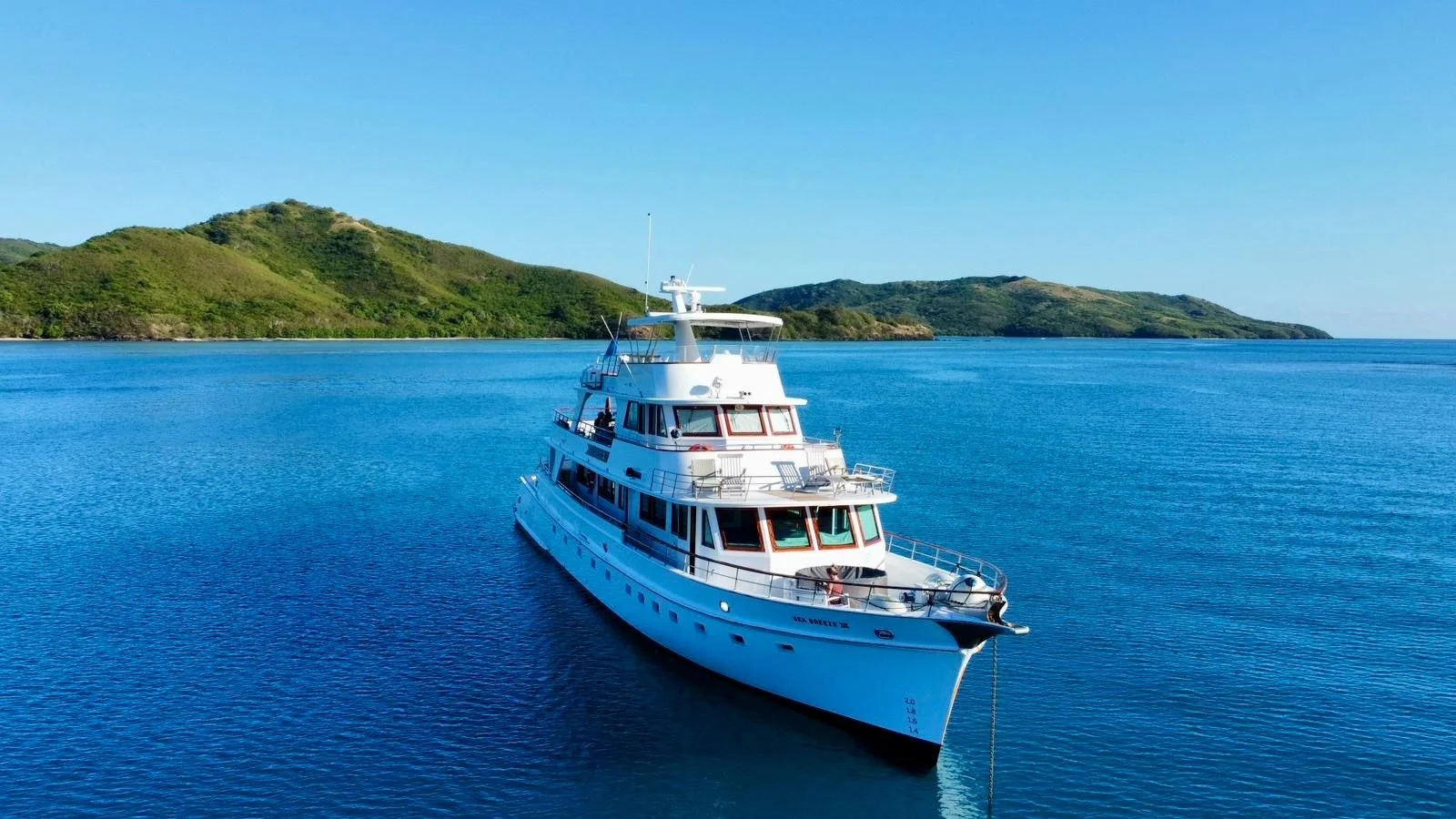 A white yacht floating on calm blue water with green hilly islands in the background under a clear blue sky.