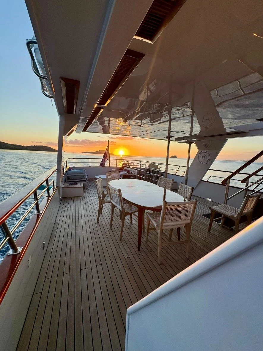 View of a yacht deck during sunset with a dining table and chairs, water, and distant islands in the background.
