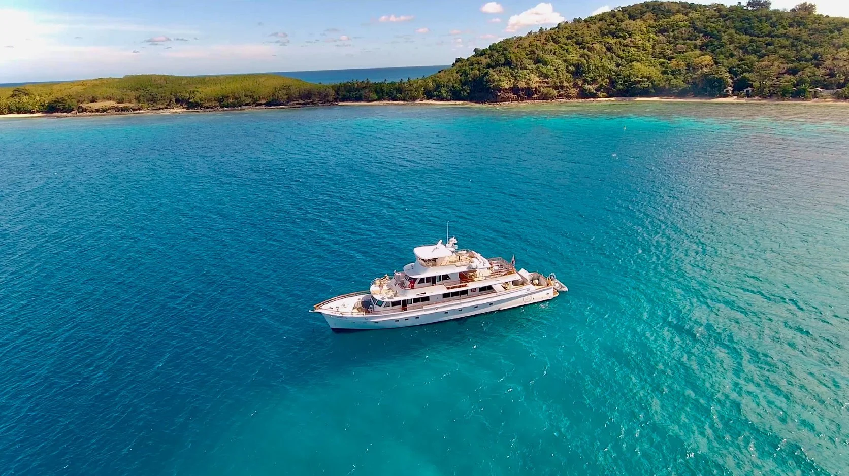 A luxury yacht on clear blue ocean water near a green island with a hilly landscape.