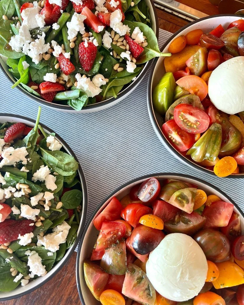 Four bowls of fresh salads and tomato salad with mozzarella cheese on a wooden table.