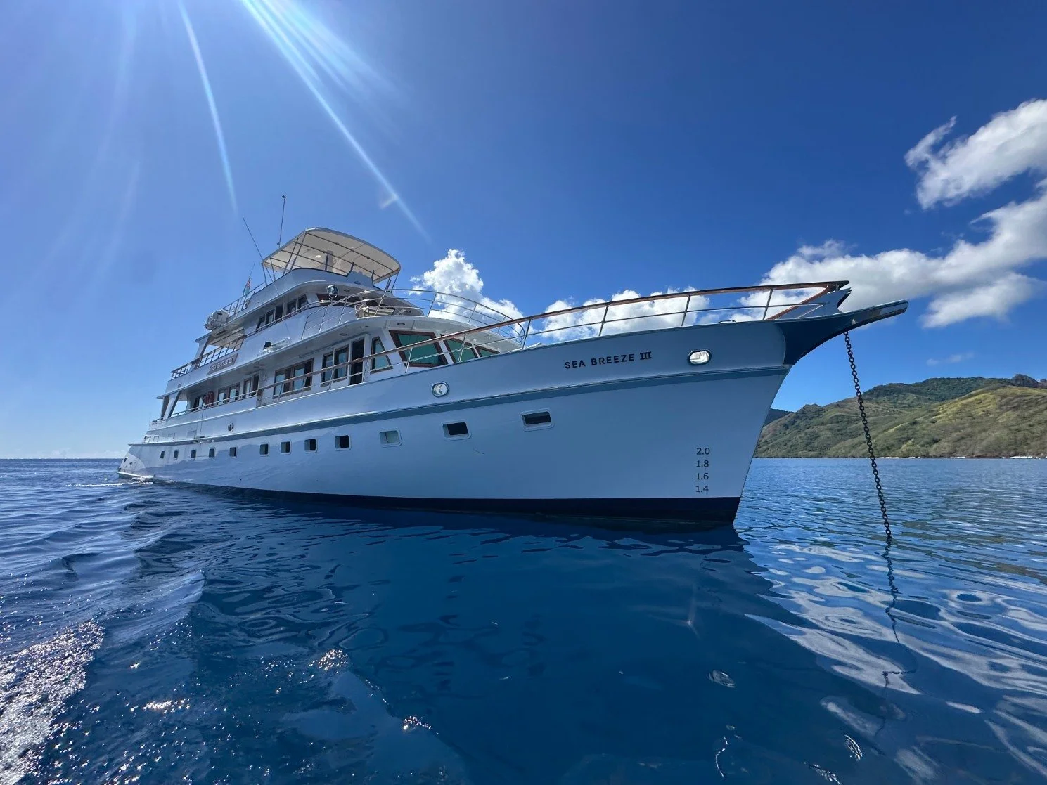 Luxury white yacht named Sea Breeze III floating on calm blue ocean waters, with a lush green island and blue sky in the background, and sunlight creating rays on the top left.