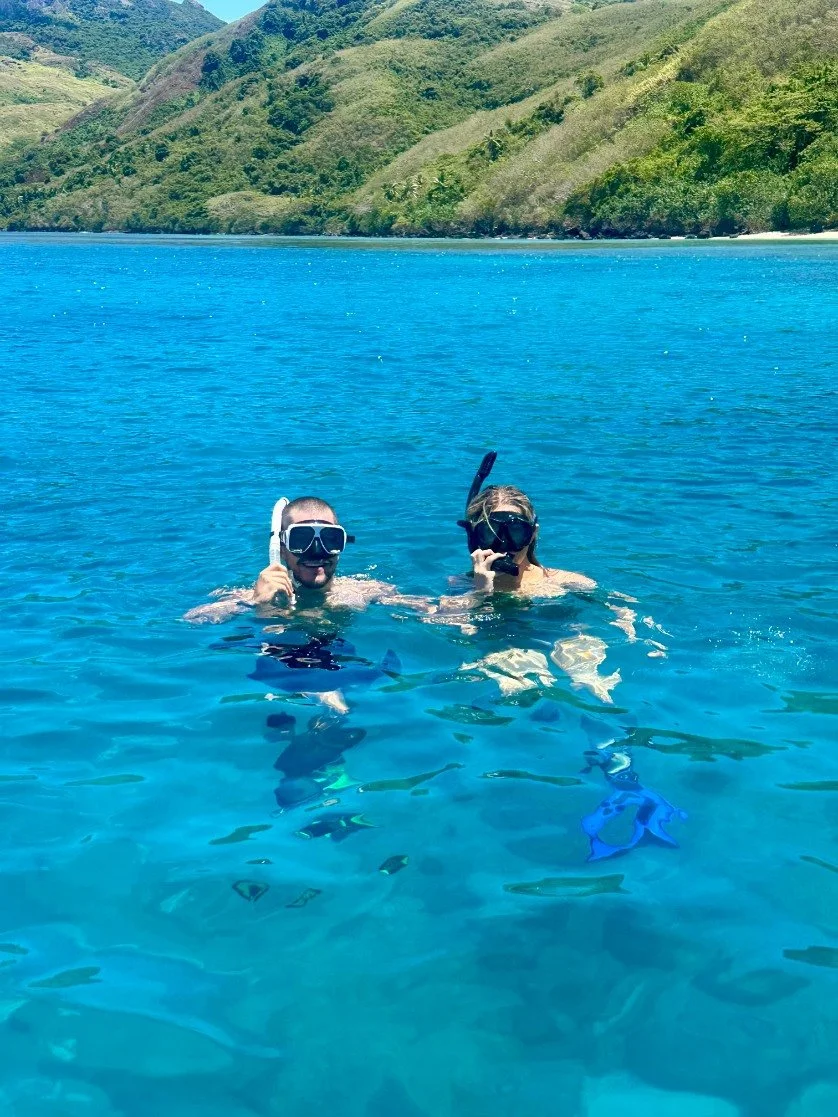 Two people snorkeling in a clear blue ocean with green hilly islands in the background.