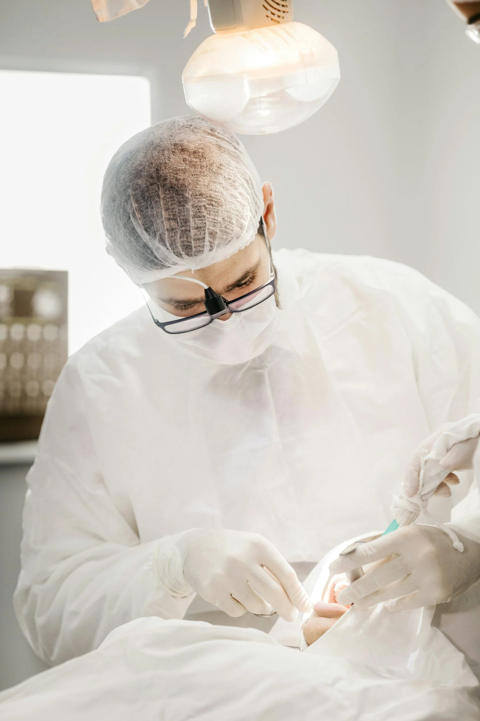 A dentist wearing a surgical mask, hairnet, and protective glasses performing a dental procedure on a patient in a well-lit clinical environment.
