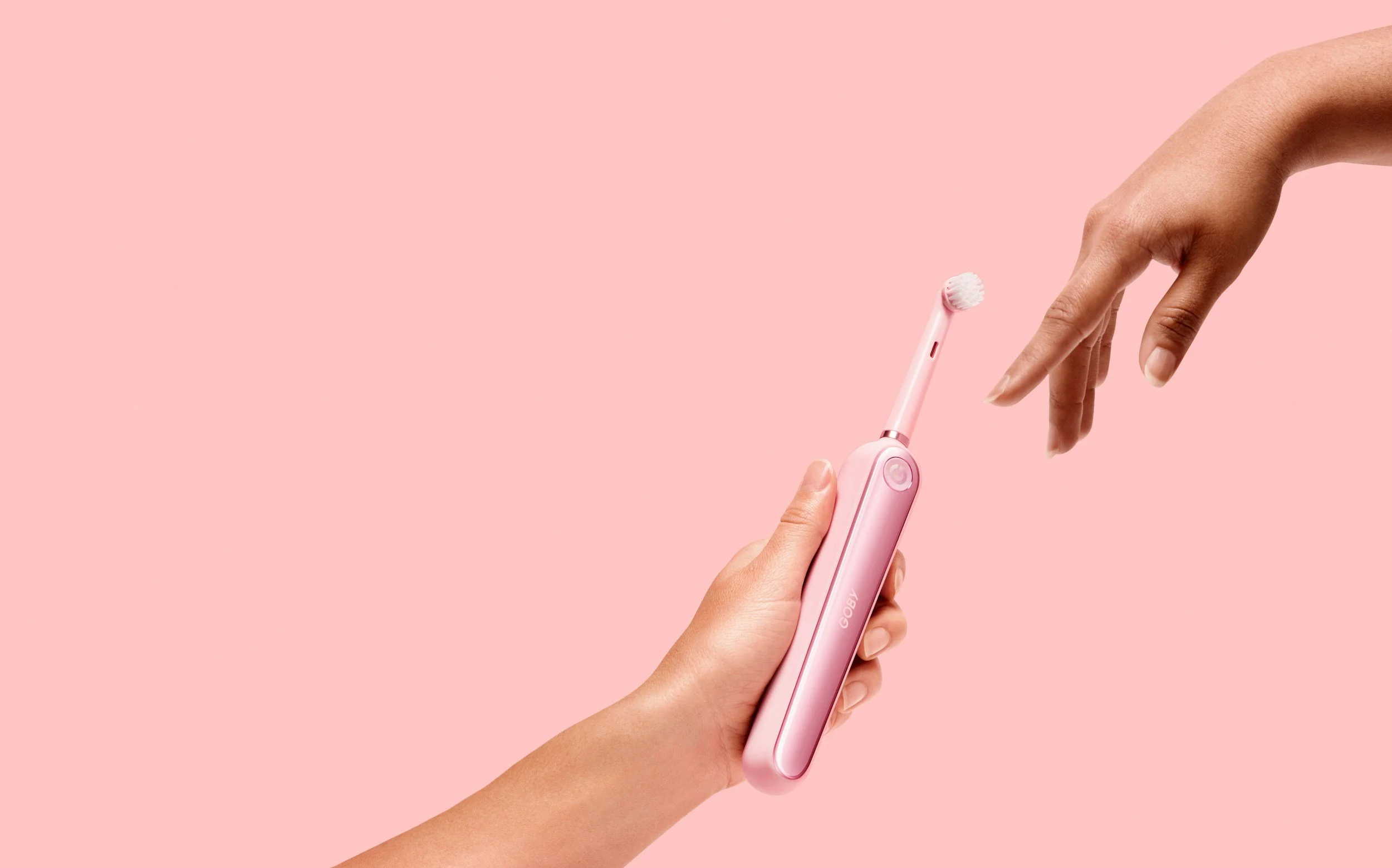 Close-up of a person's hand holding a pink GOBY electric toothbrush, with another person's hand reaching toward it against a pink background.
