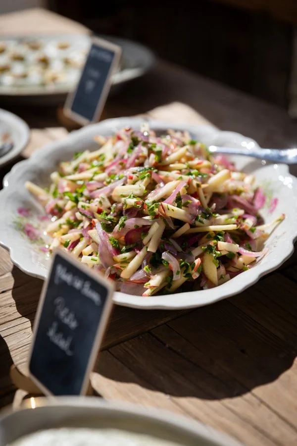 A bowl of apple and radish salad garnished with herbs on a wooden table, with a small sign and a spoon nearby.