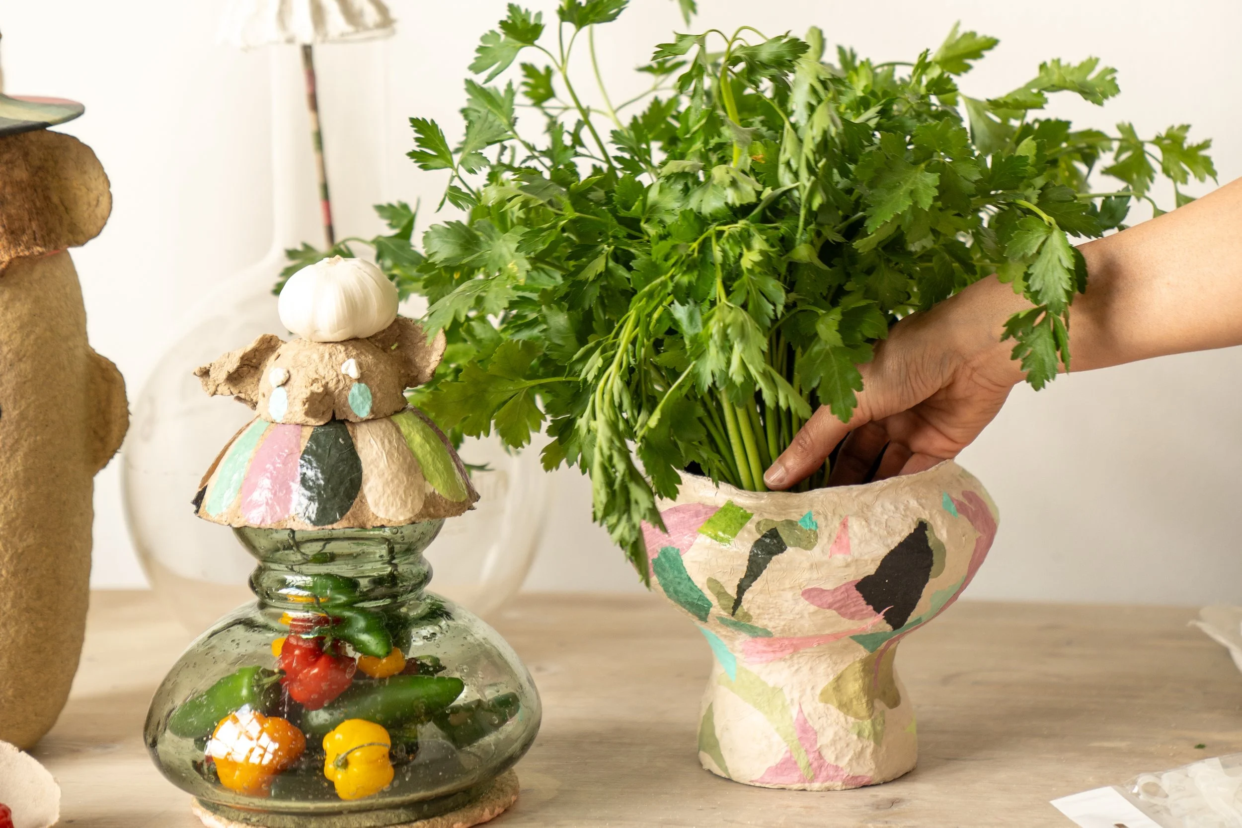A person arranging a bouquet of fresh cilantro in a colorful, decorated paper mache vase. There are additional decorative items such as a sculpture and a glass container filled with small vegetables on a wooden surface.
