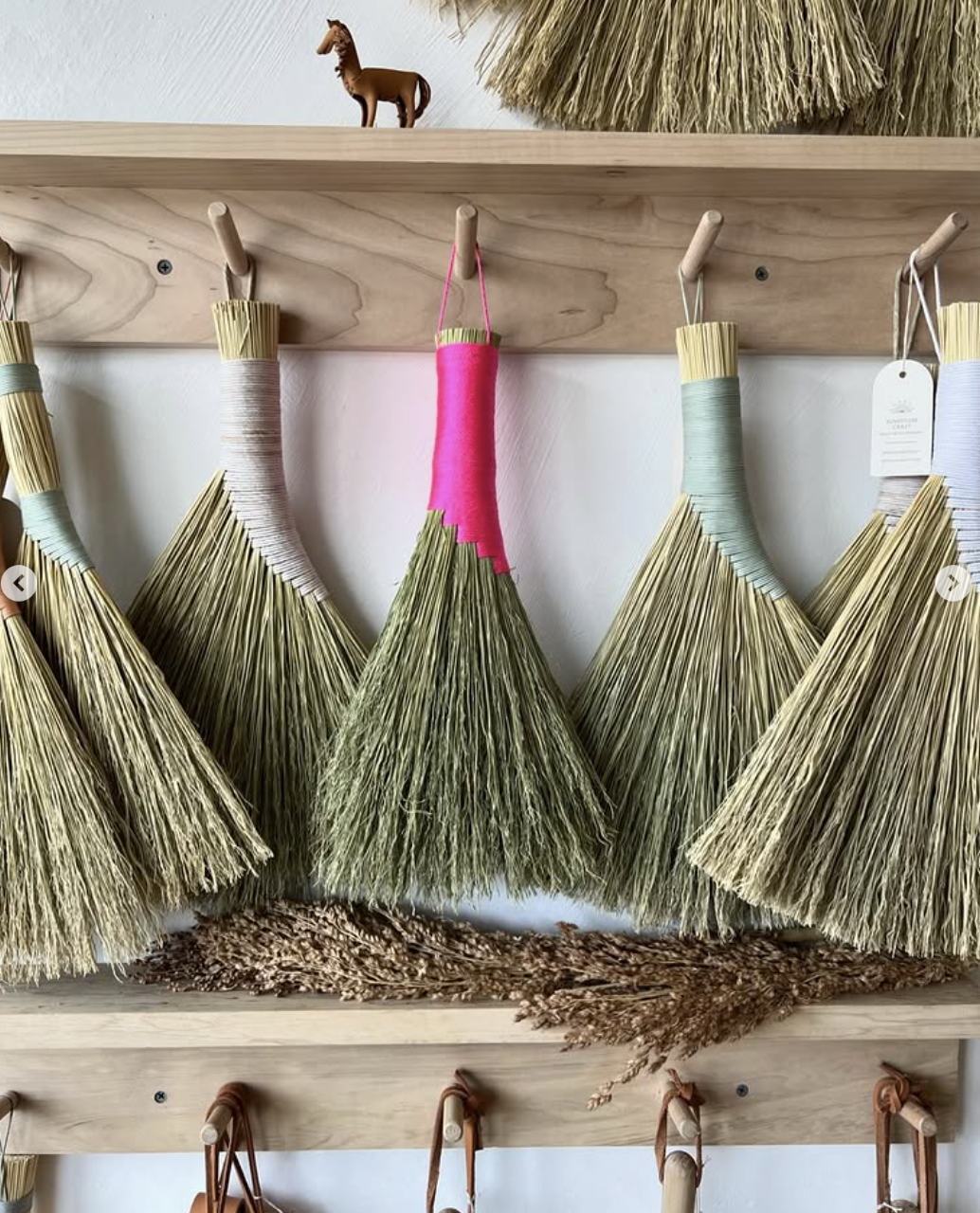 Colorful straw broom heads hanging on a wood rack with a small wooden horse figurine on a shelf above.