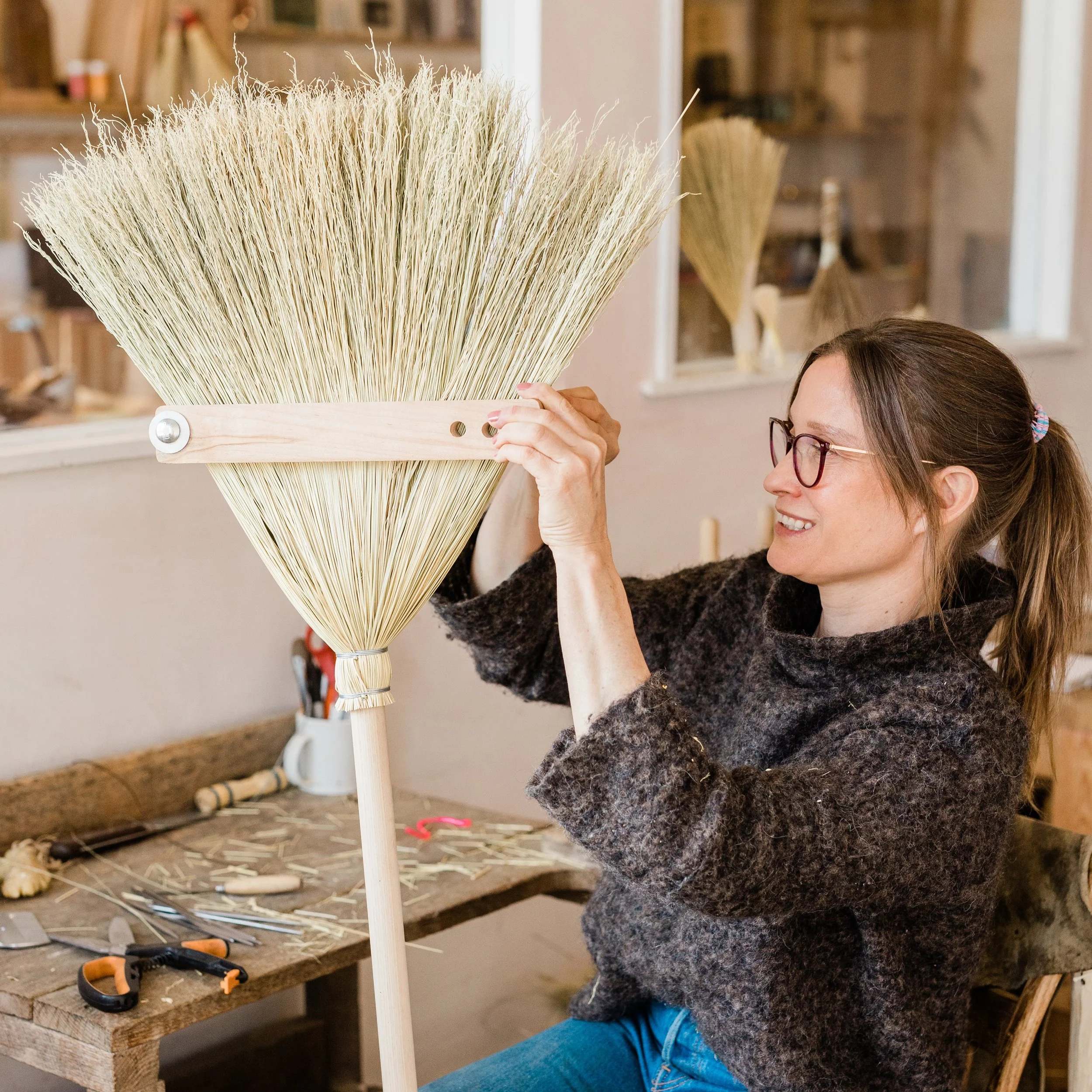 A woman is working on a woodworking project creating a large broom with a wooden handle and straw bristles in a workshop.